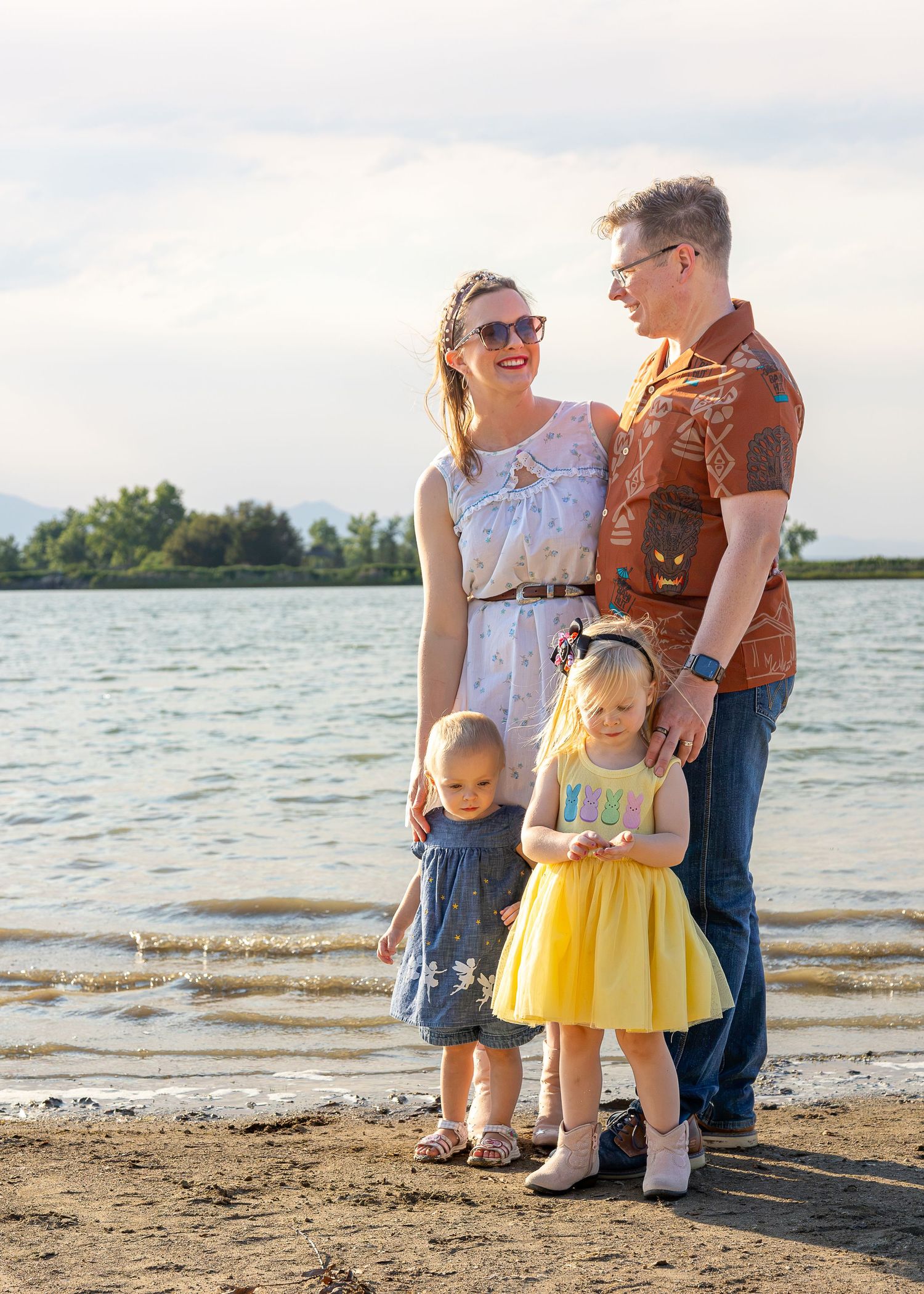 Family of four posing by a lakeside, parents smiling, two young girls in colorful dresses on a sunny day.