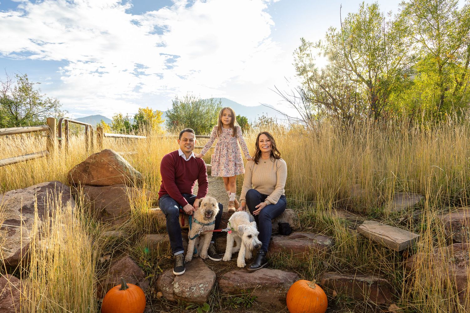 Family of three with two white dogs sitting on stone steps surrounded by autumn foliage and pumpkins outdoors.