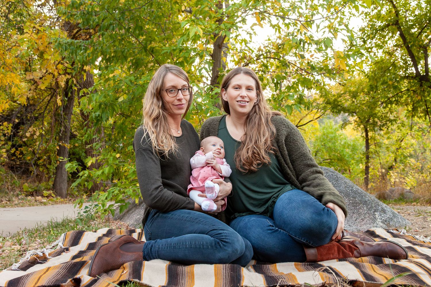Two women sitting on a blanket outdoors in autumn, one holding a newborn baby, surrounded by colorful fall foliage.
