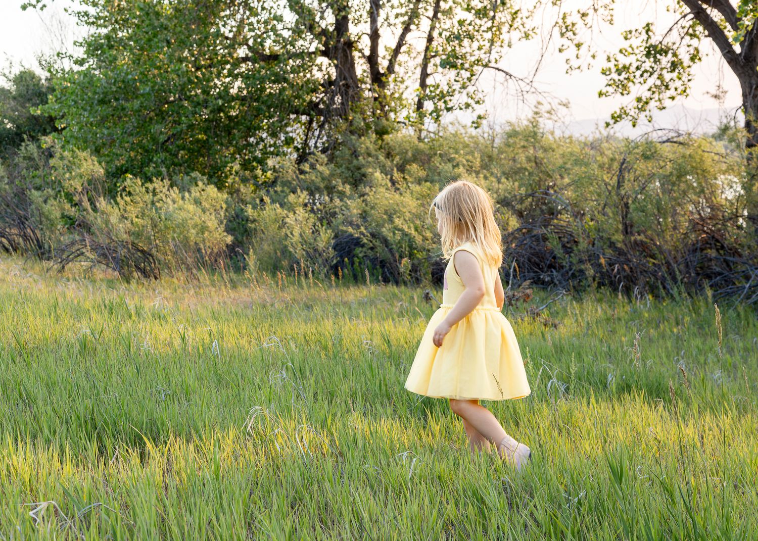 A young girl in a yellow dress walks barefoot through a lush green meadow surrounded by trees.