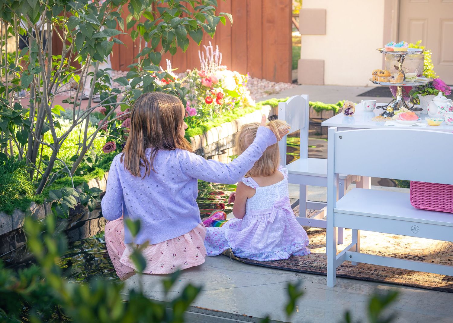 Two young girls in pastel dresses sit by a garden path beside a white outdoor table, surrounded by lush green plants and colorful flowers.