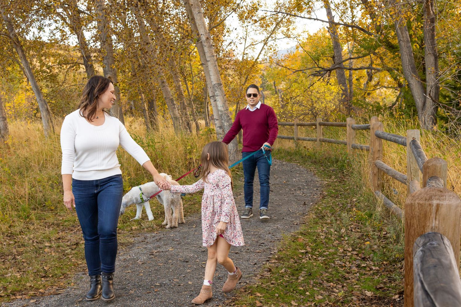 Family walking on a scenic autumn path with a dog, surrounded by golden trees and a wooden fence.