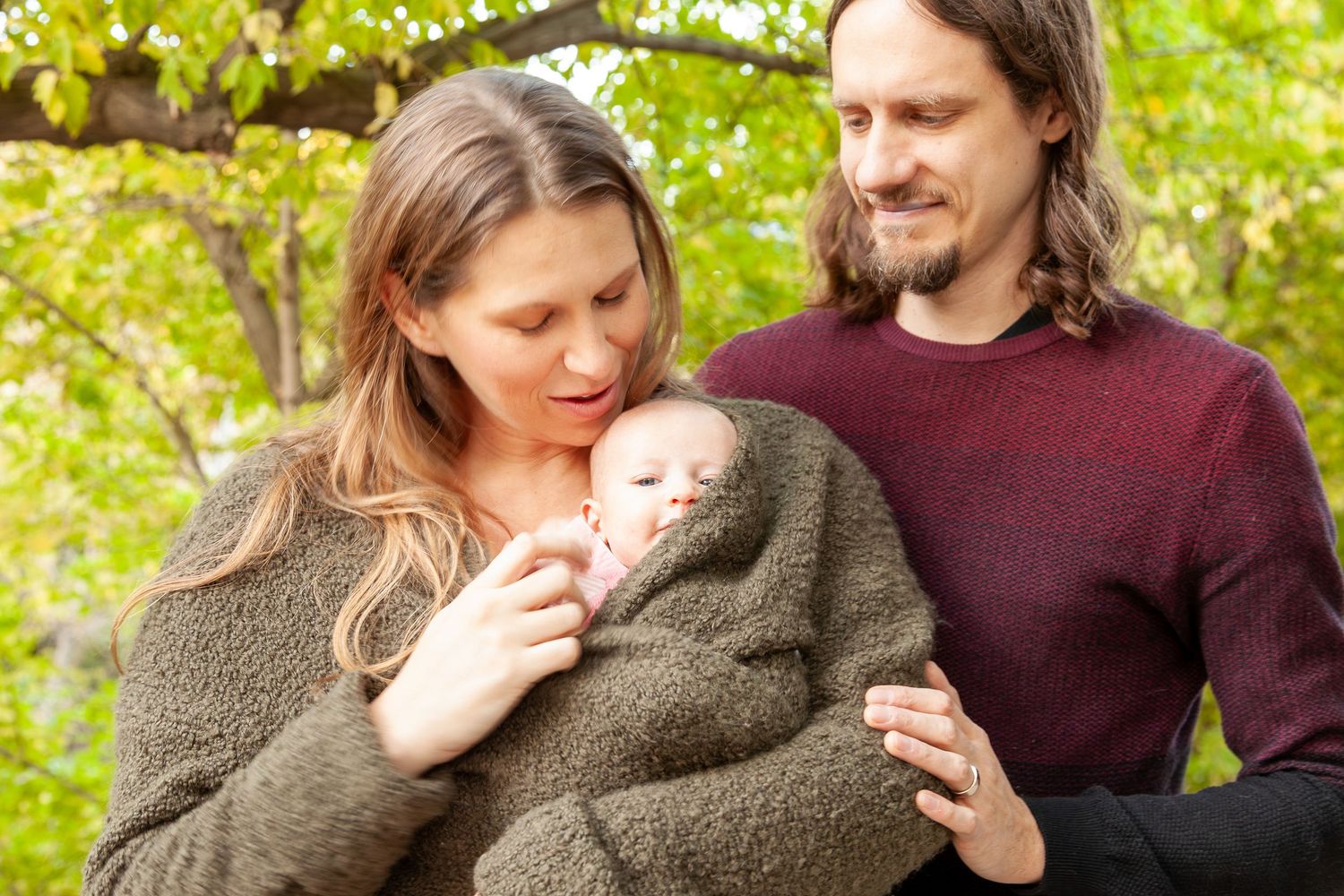 A couple outdoors admiring their newborn baby wrapped in a cozy green blanket among autumn trees.
