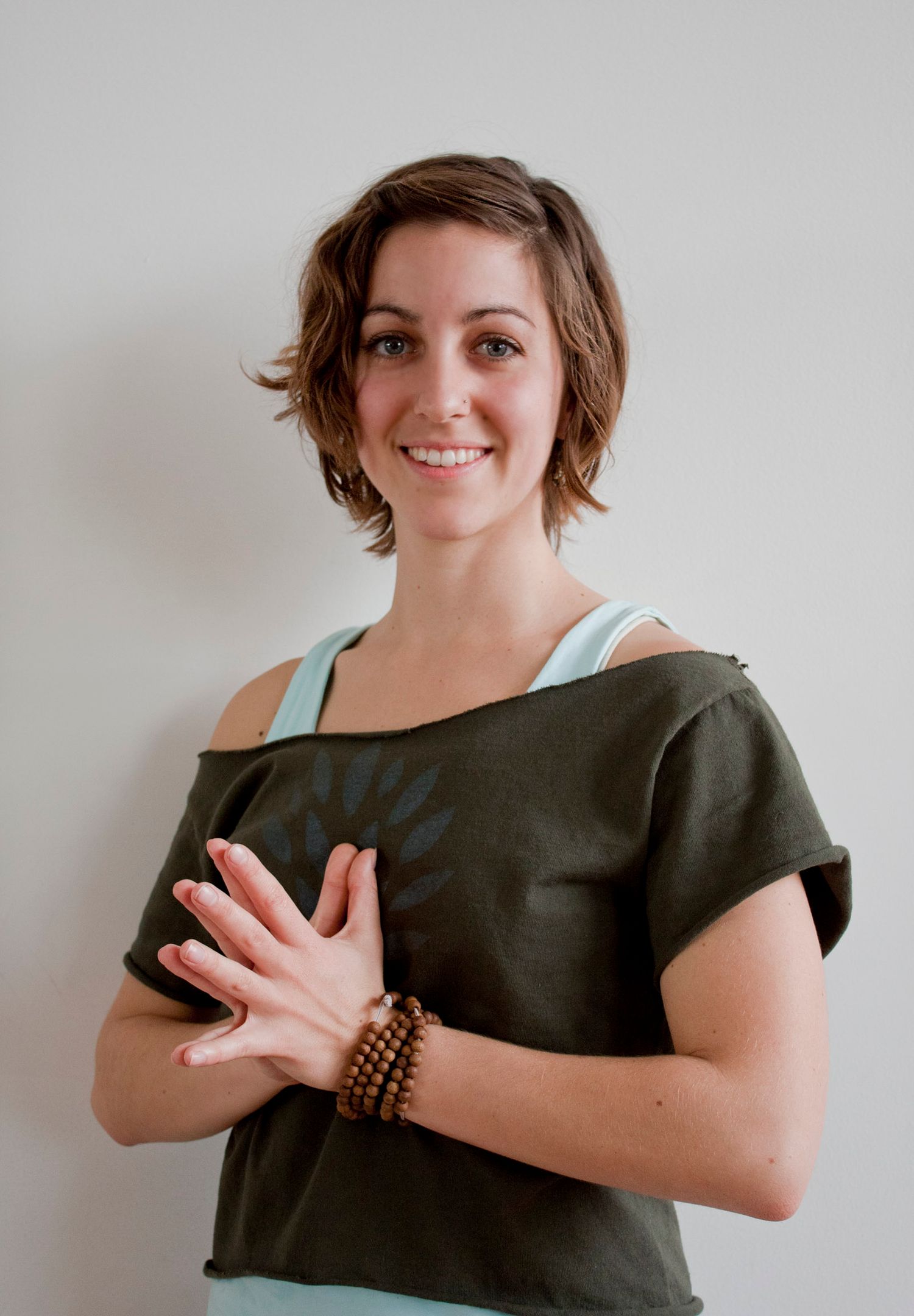 Smiling brunette woman in olive green top with prayer hands gesture wearing wooden bracelet.