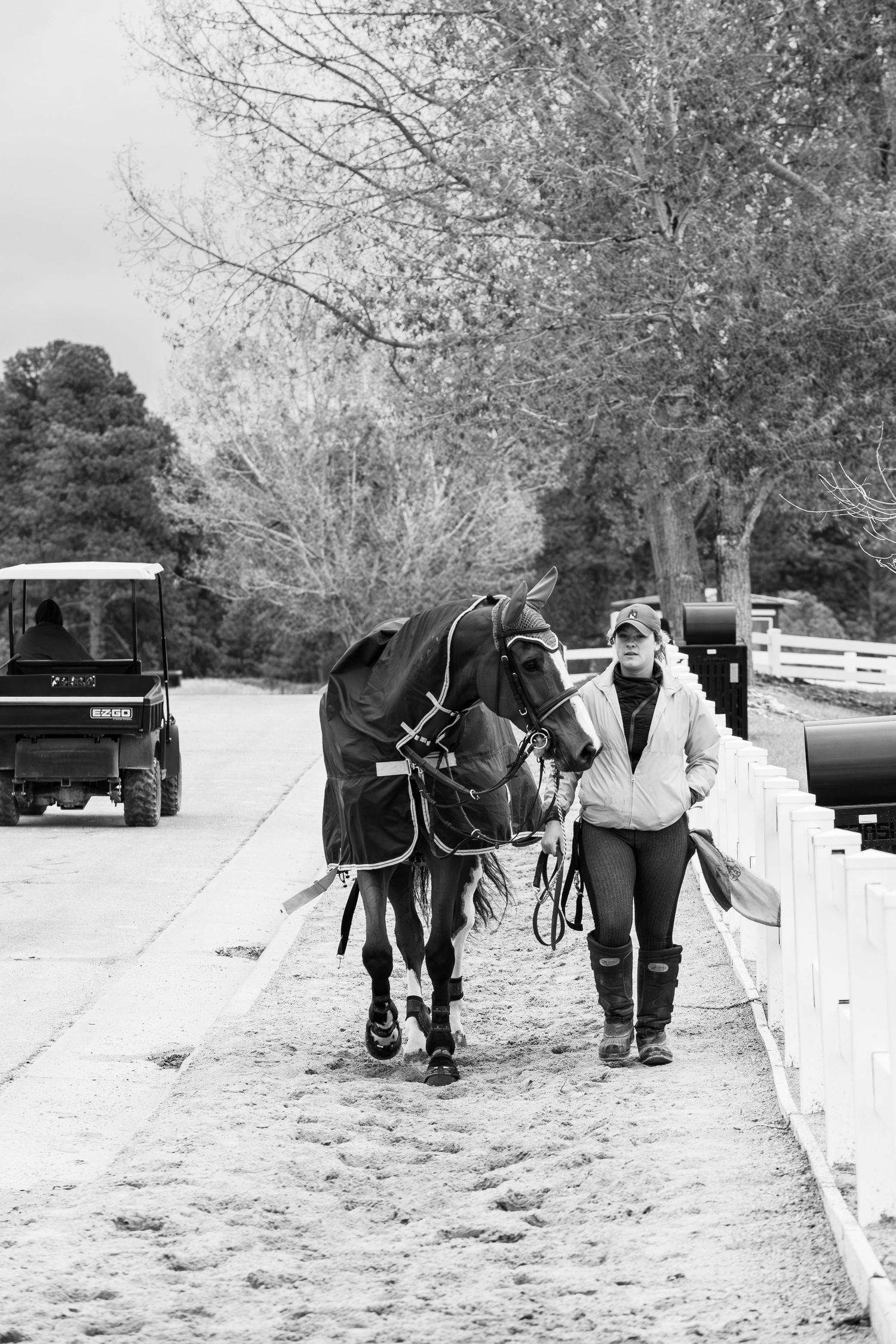 Woman walking a blanketed horse along a white fence path with a golf cart in background.