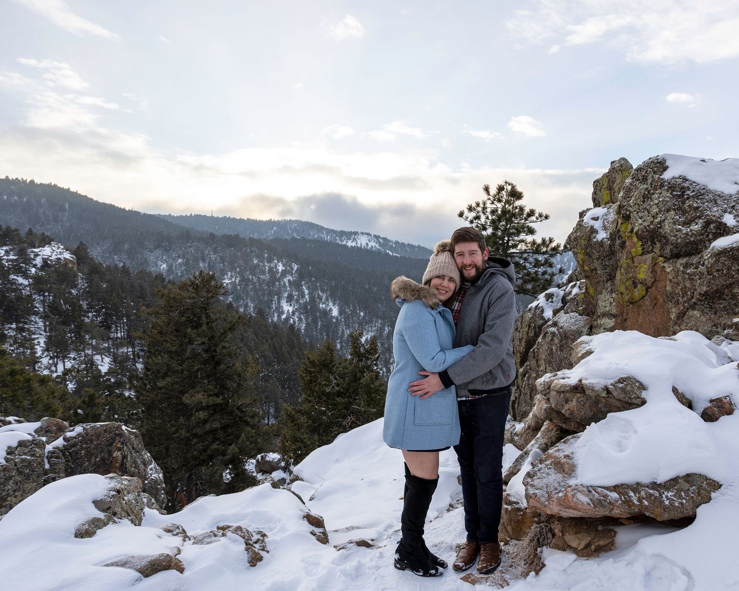 Happy couple embracing on snowy mountain overlook with scenic winter landscape behind them.