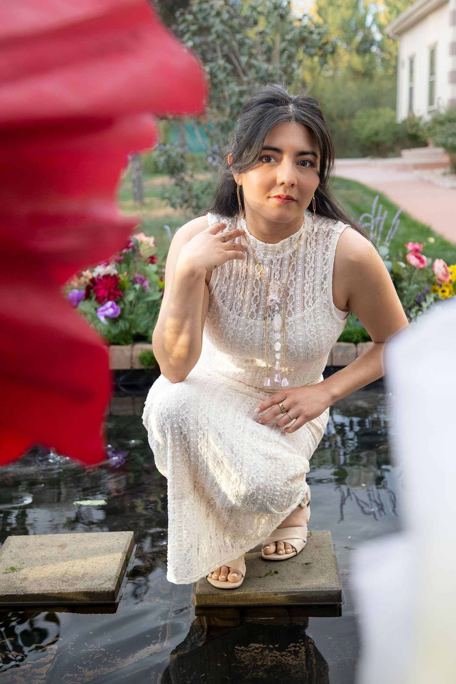Woman in a cream lace dress crouches on stepping stones over a garden pond, with colorful flowers and a red blurred petal in the foreground.