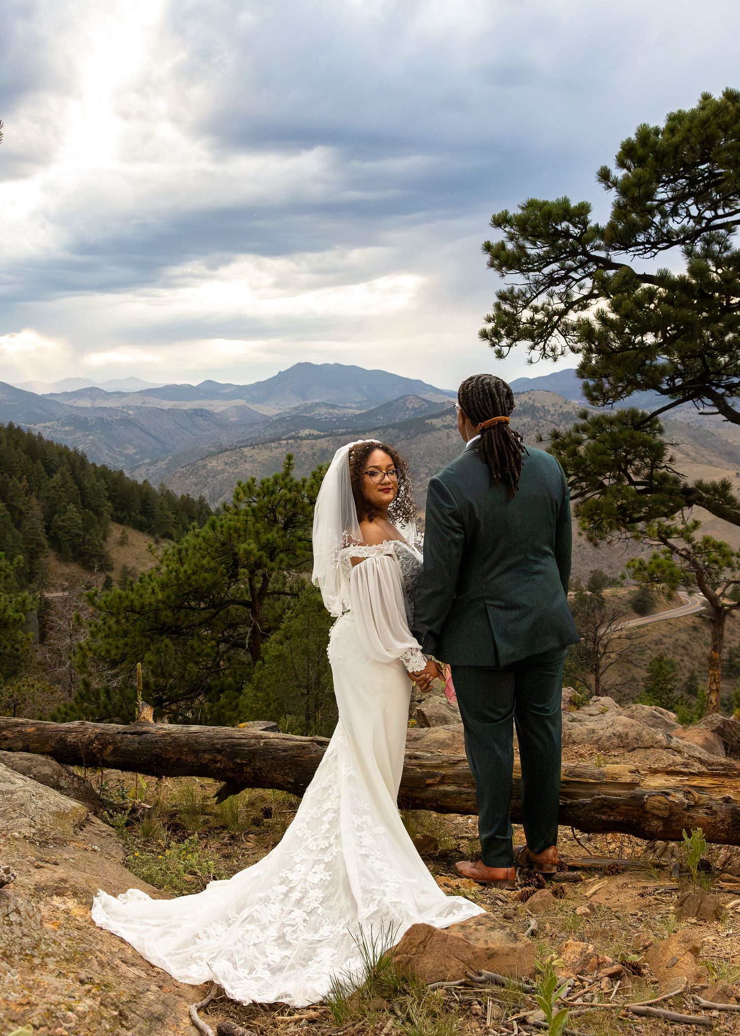 A bride and groom hold hands at a mountain overlook, with stunning Rocky Mountain views and dramatic clouds behind them.