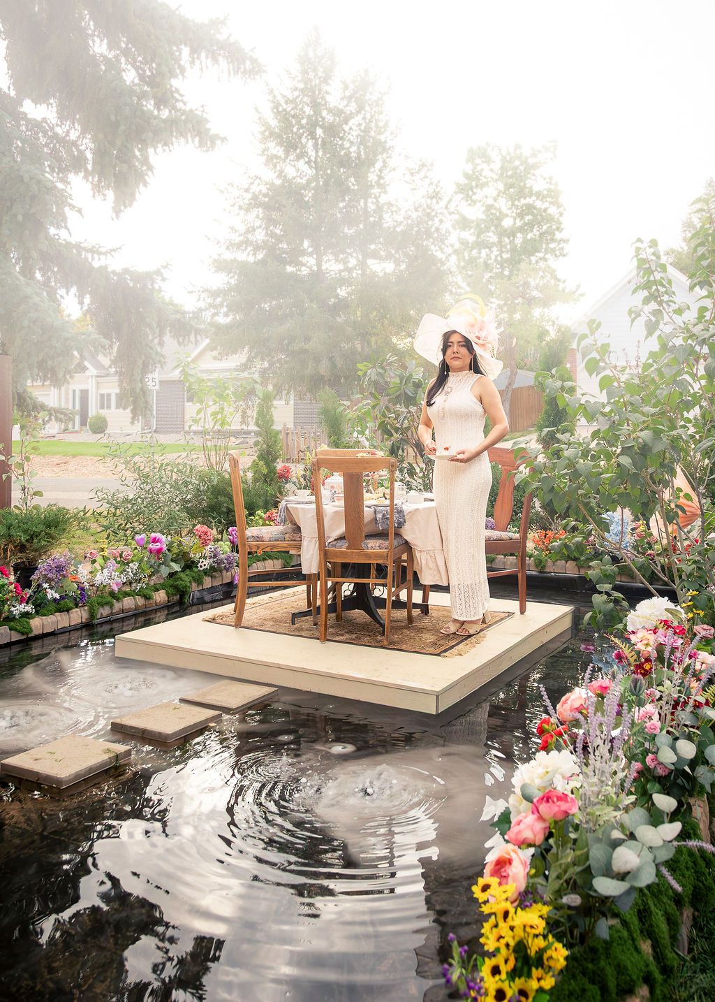 Woman in a white dress and wide-brimmed hat stands beside a dining table on a platform over a pond, surrounded by colorful flowers.