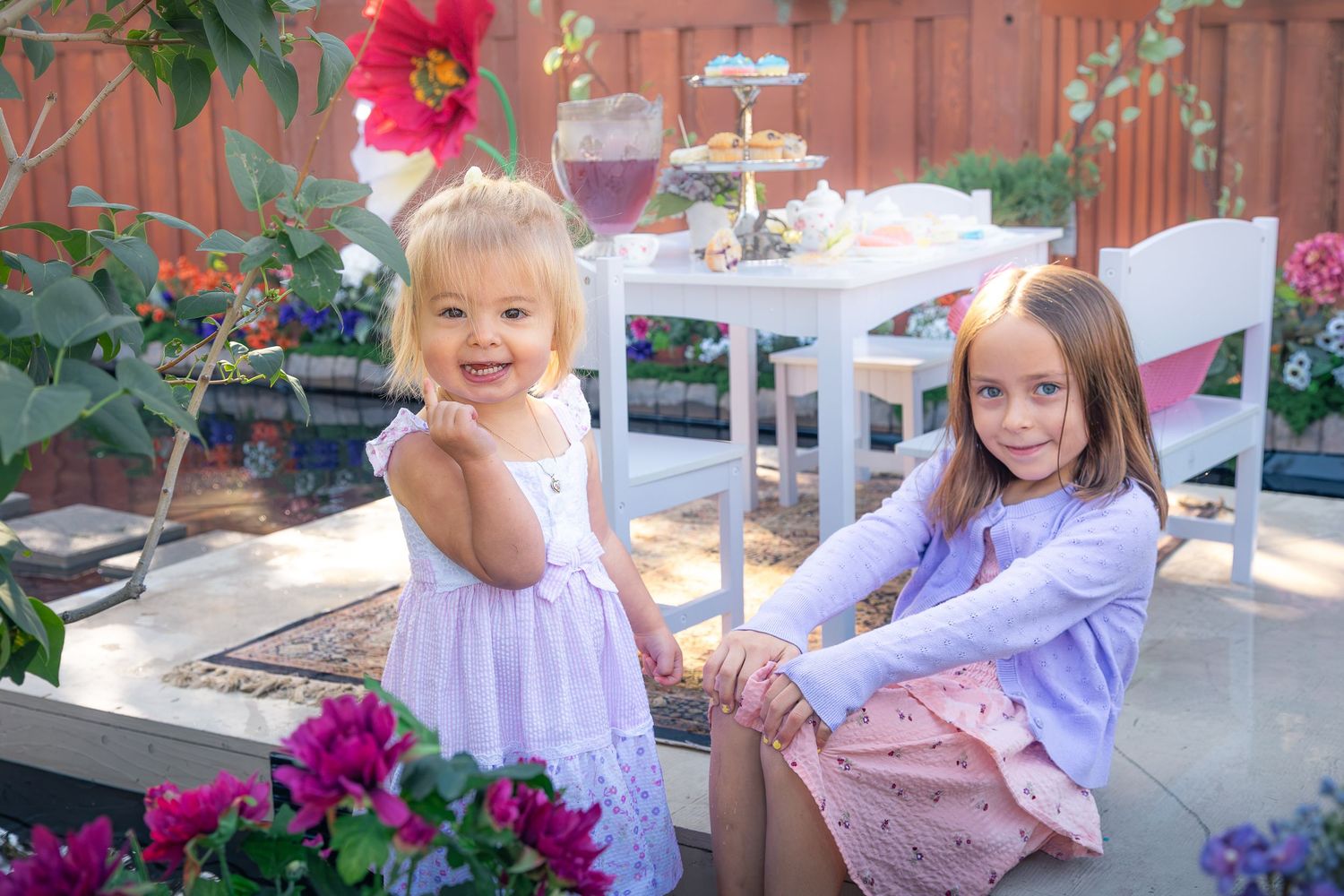 Two young girls in pastel dresses sit on a sunny patio near a white table with a tea party spread, surrounded by colorful flowers.