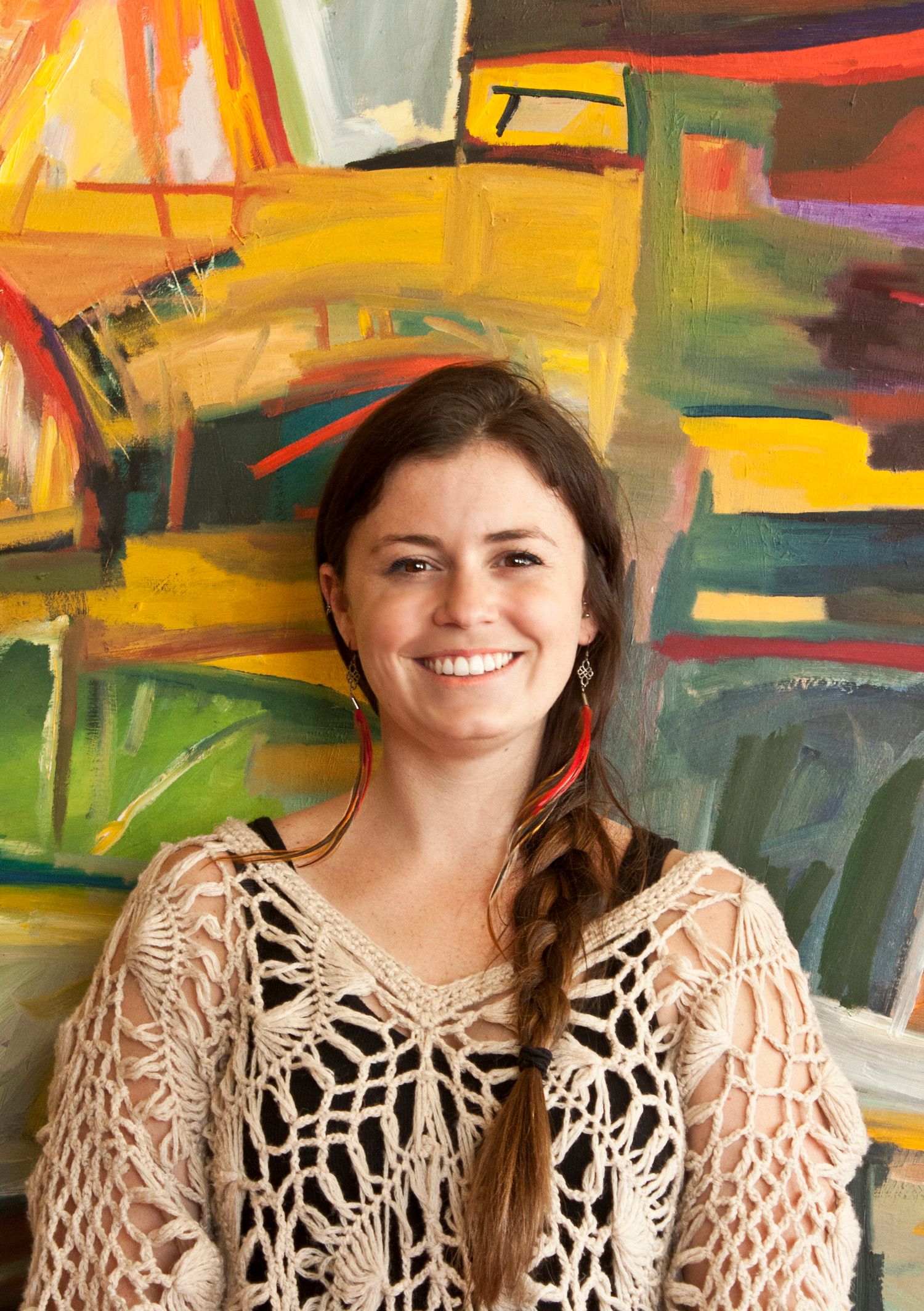 Smiling young woman with braided hair wearing crochet top standing before a vibrant colorful abstract painting.