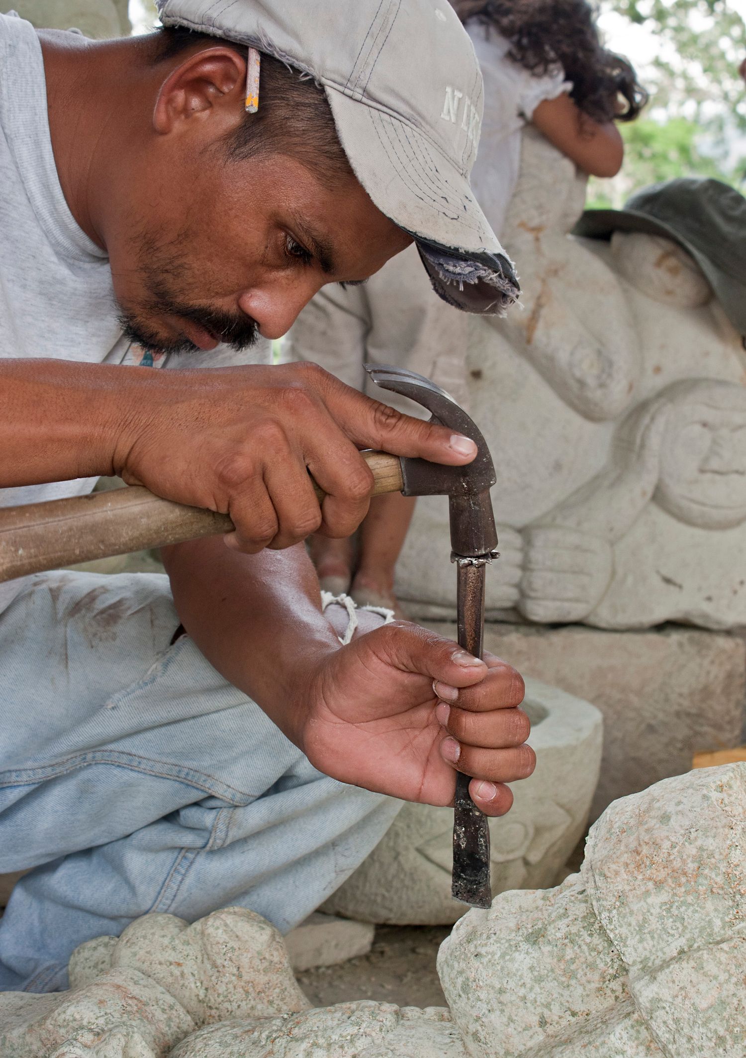 A craftsman carefully chisels a stone sculpture outdoors surrounded by carved stone figures.