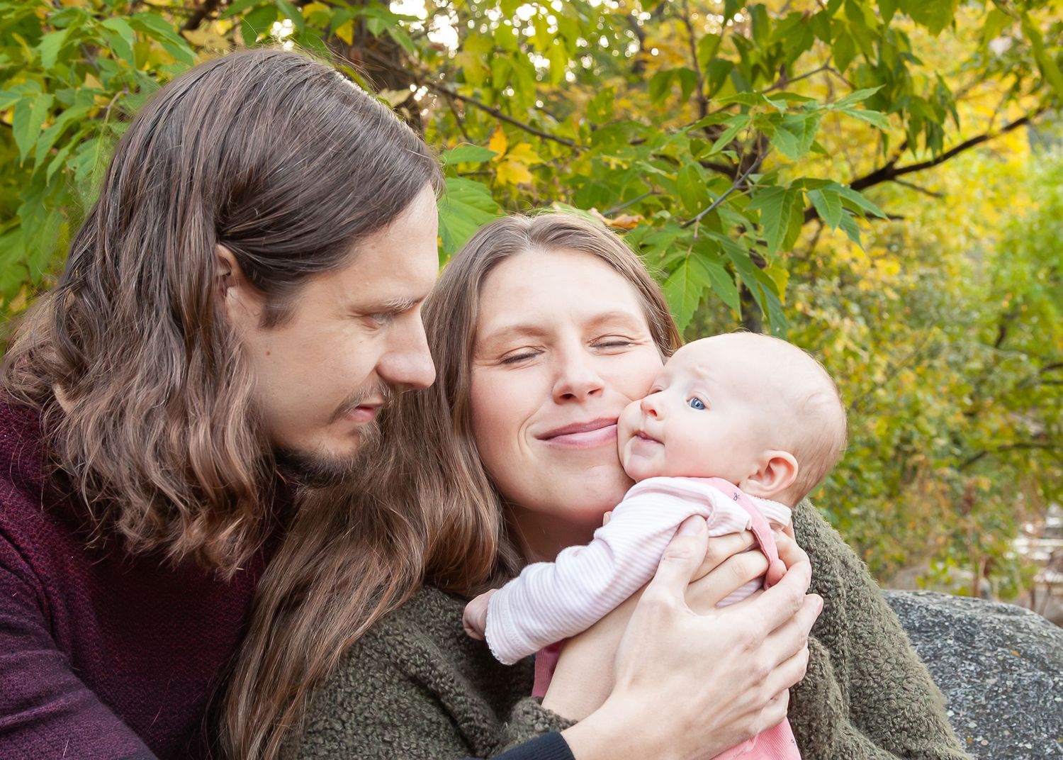 Young couple with long hair tenderly holding and smiling at their newborn baby outdoors in autumn.