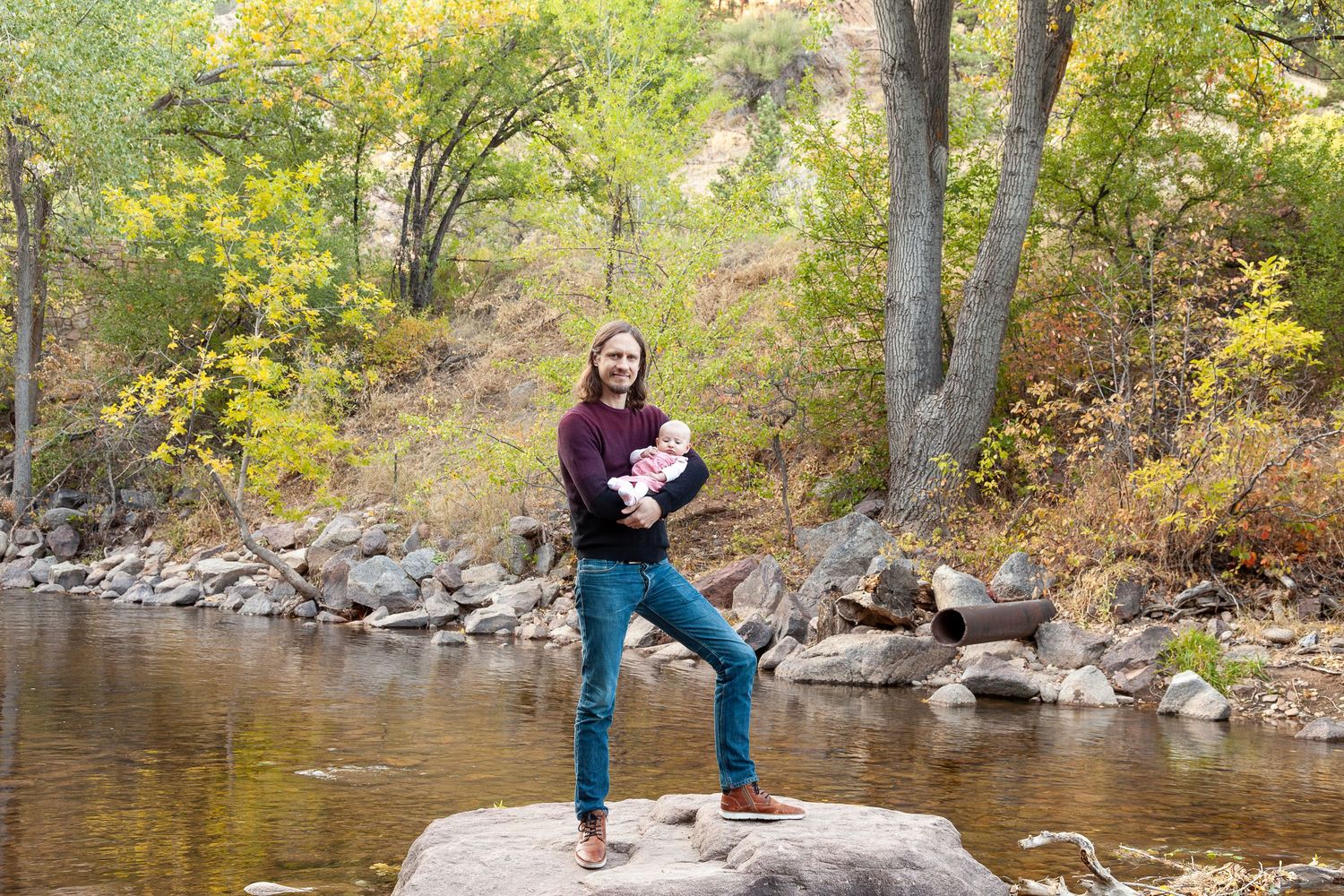 A man holding a baby stands on a rock by a peaceful river surrounded by autumn trees in a scenic outdoor setting.