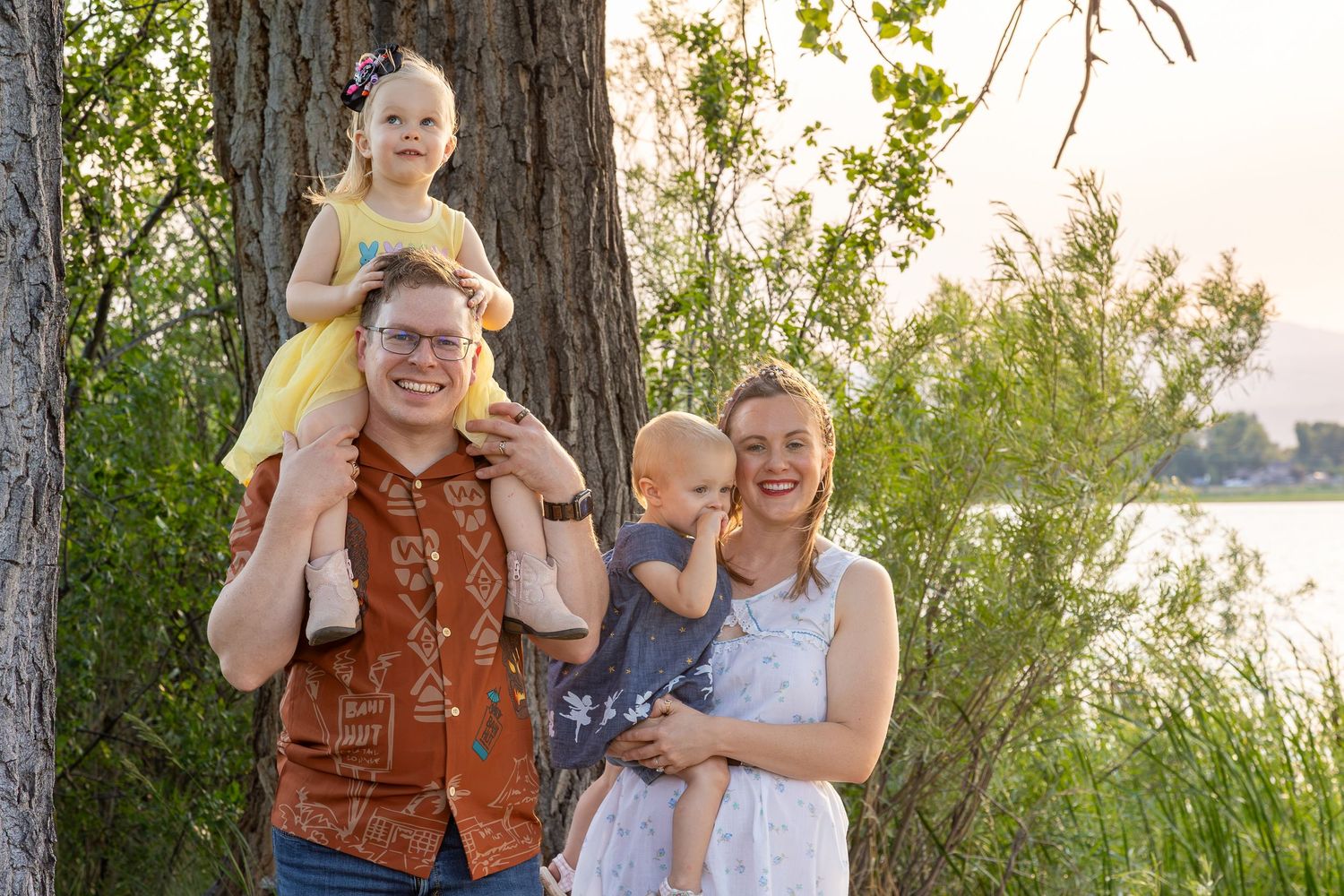 Family of four posing outdoors near a tree by a lake, with two young children, on a warm sunny day.