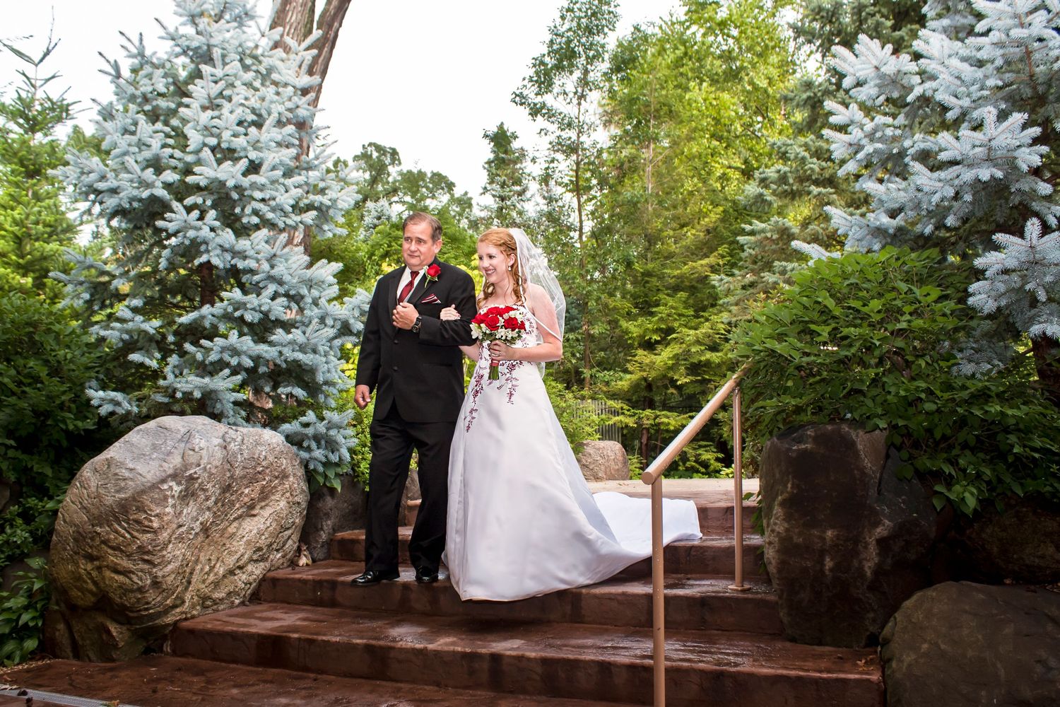 Bride and groom descend stone steps surrounded by lush evergreen trees in an outdoor wedding ceremony.