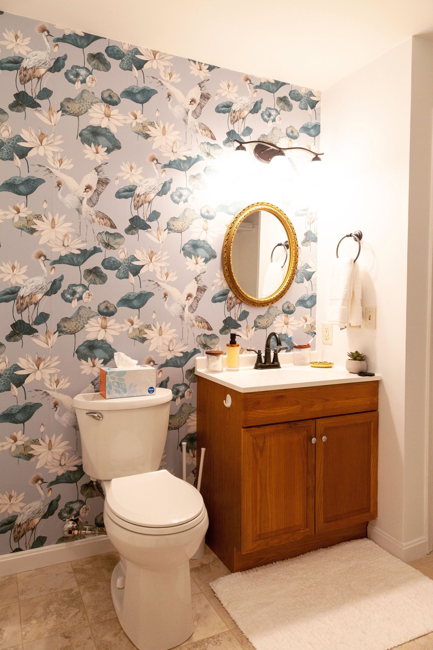 Bathroom with floral wallpaper, wooden vanity, gold oval mirror, and white toilet on beige tile floor.