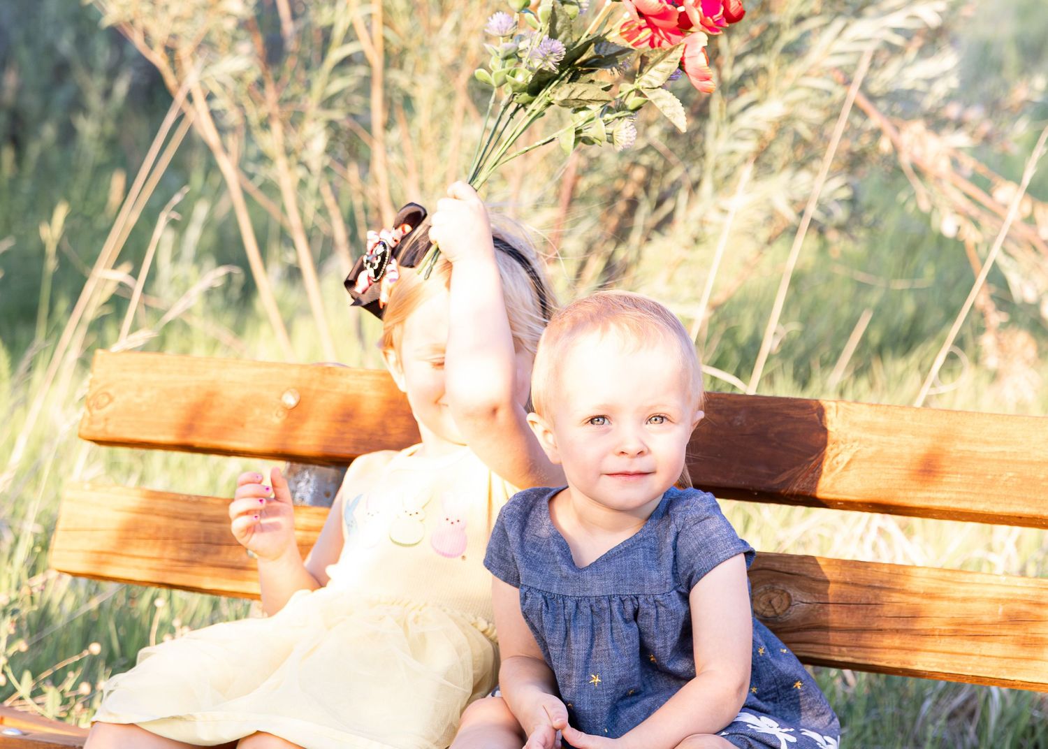 Two young sisters sitting on a wooden bench outdoors, one holding a bouquet of wildflowers.