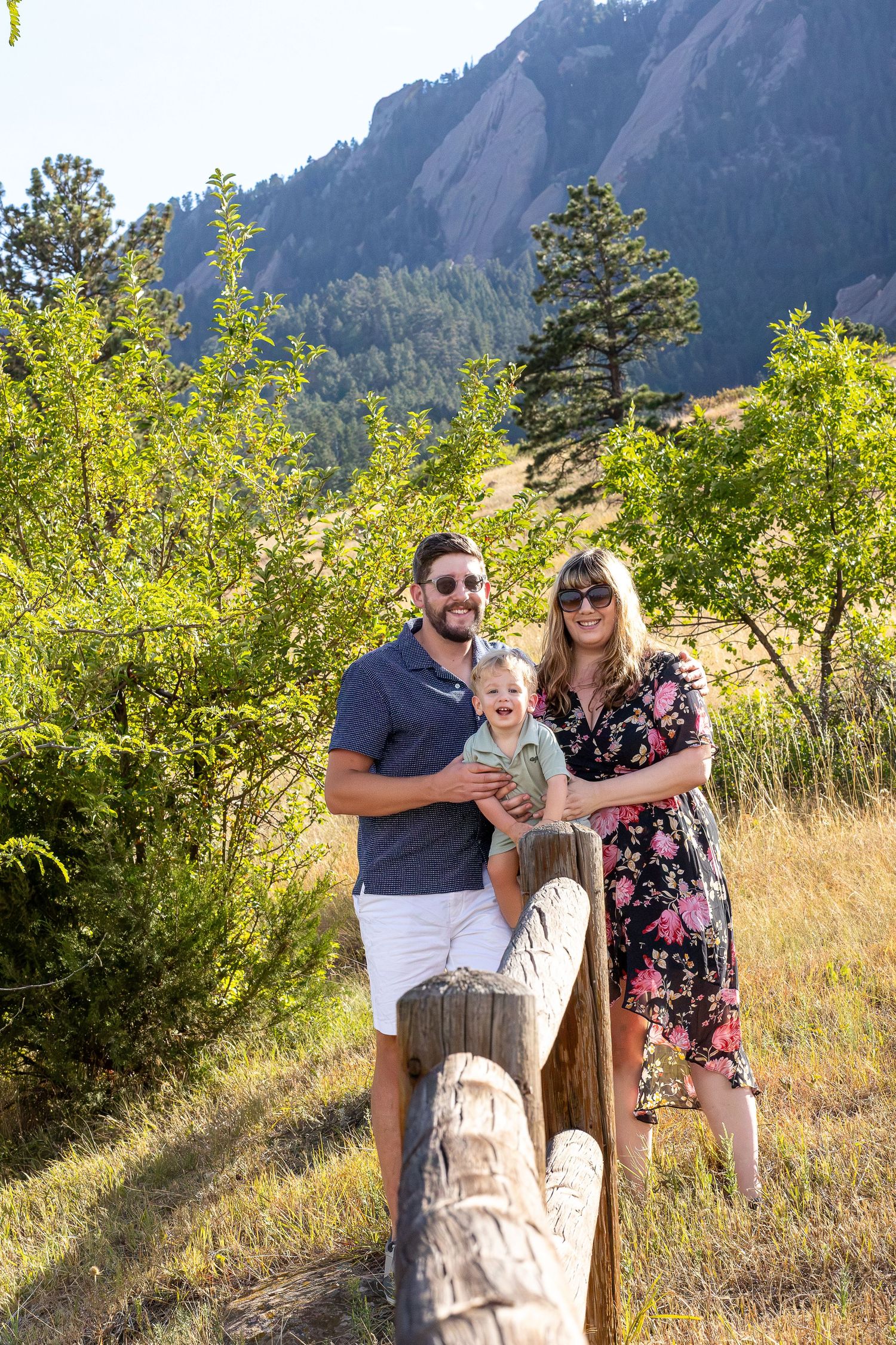 A smiling family with a baby poses outdoors near wooden posts with lush greenery and rocky Flatirons mountains in Boulder, CO.