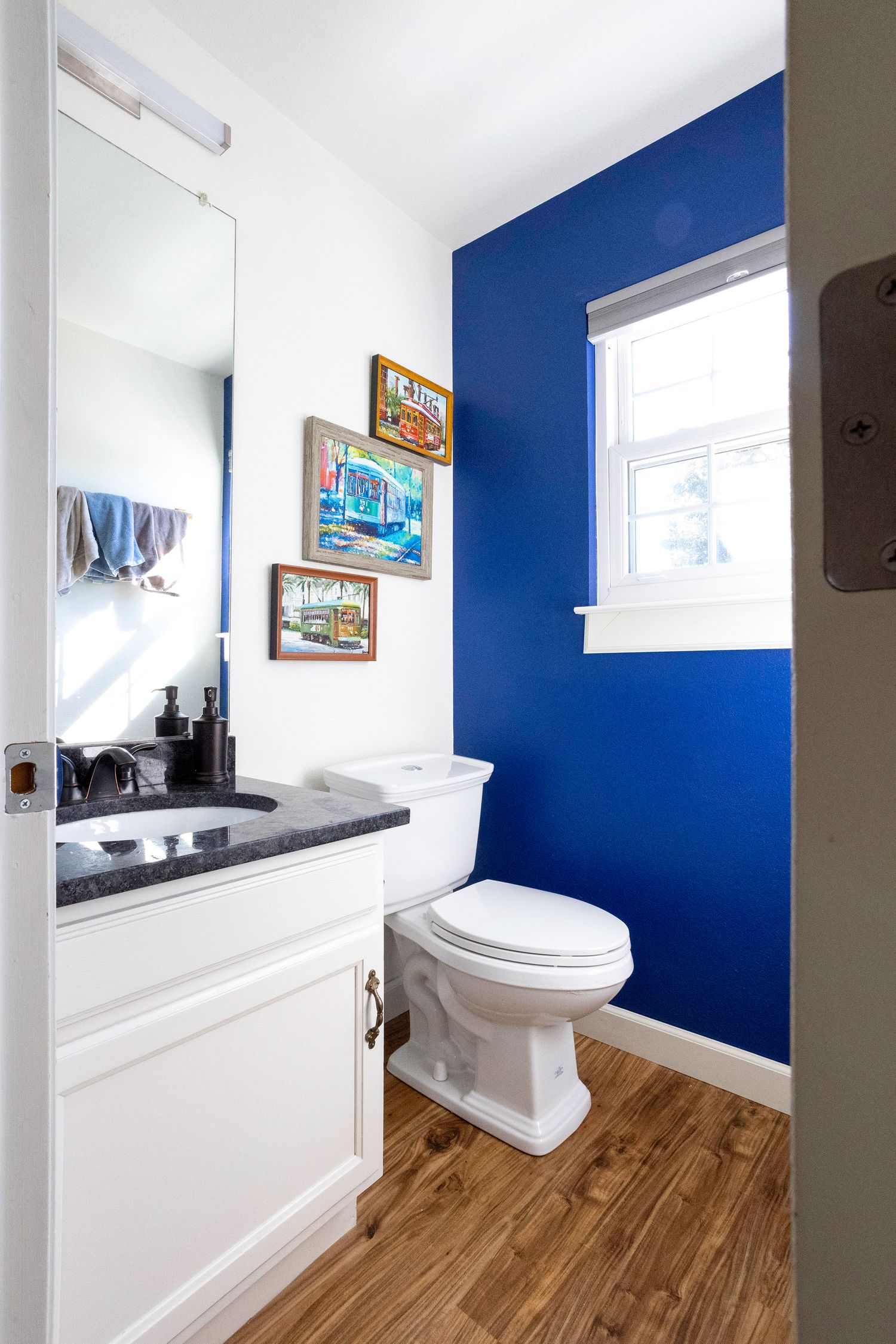 Half bathroom with blue accent wall, white vanity, dark countertop, wood floors, and framed artwork.
