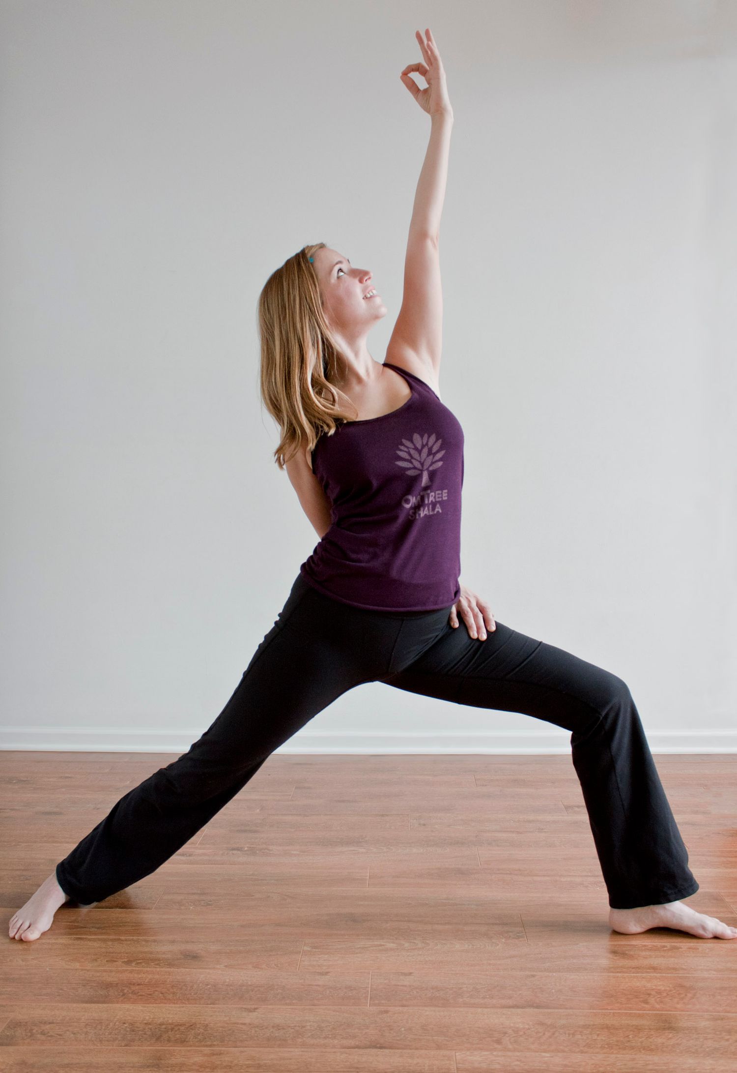 Blonde woman in purple OmTree Shala tank performing Warrior I yoga pose on hardwood floor.