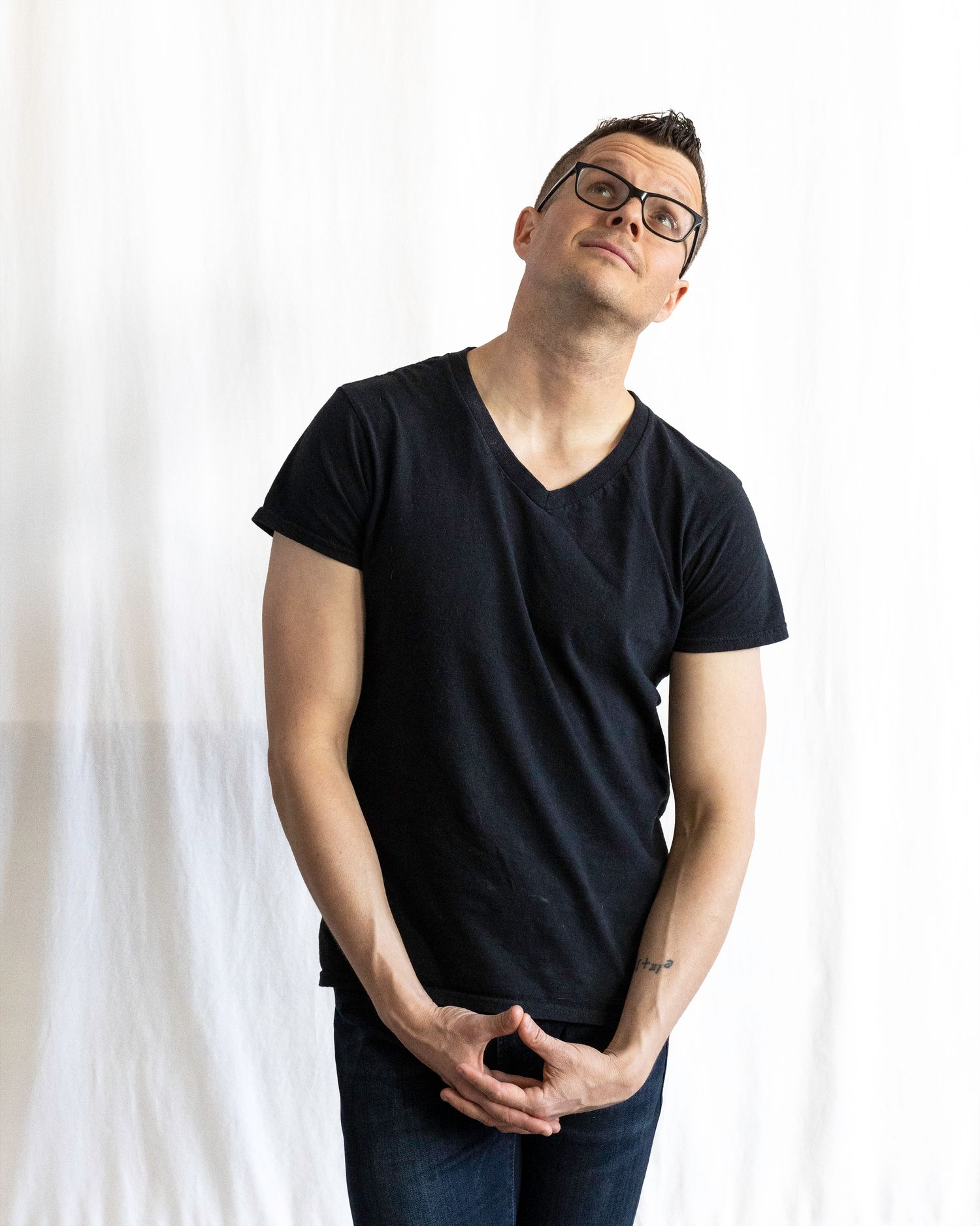 Man in black v-neck t-shirt and jeans looking upward while standing against a white curtain.