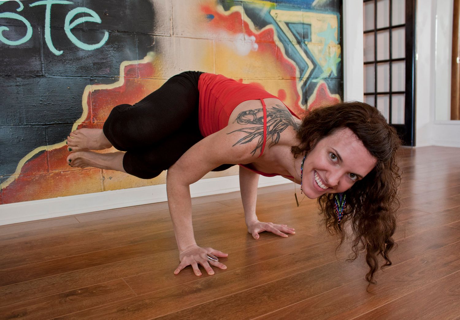 Woman with curly hair and tattoos performing crane yoga pose on wooden floor near graffiti wall.