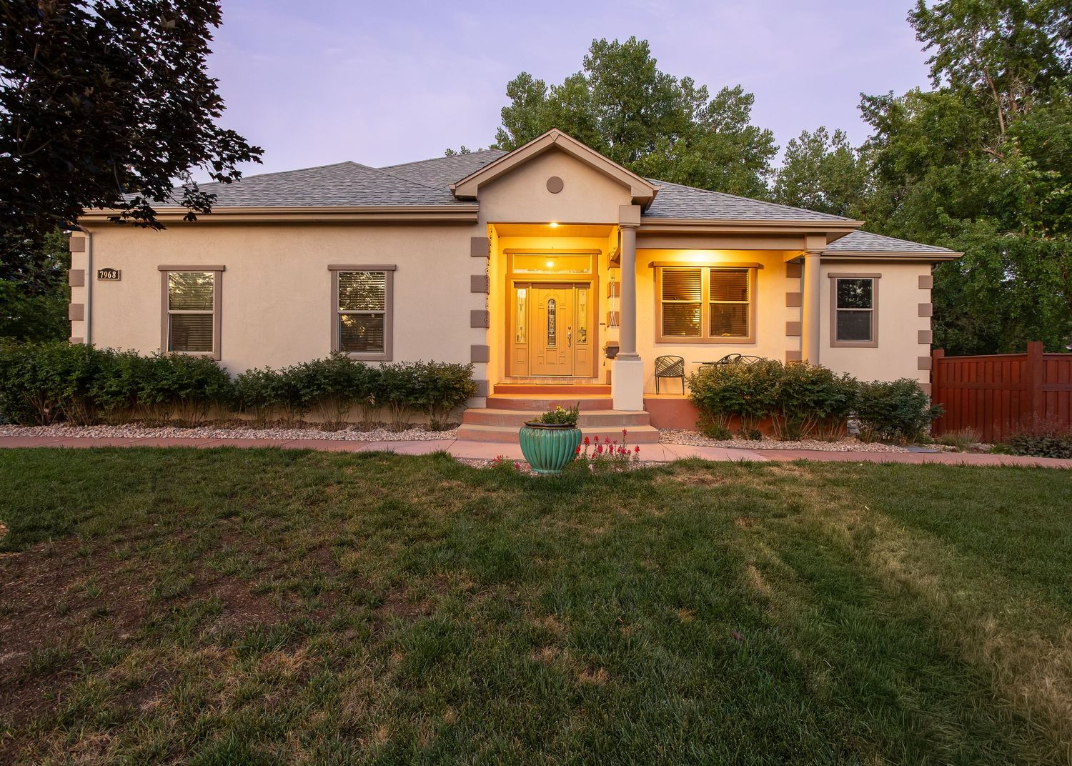 Charming stucco ranch home with warm lit entryway, lush green lawn, and mature trees at dusk.