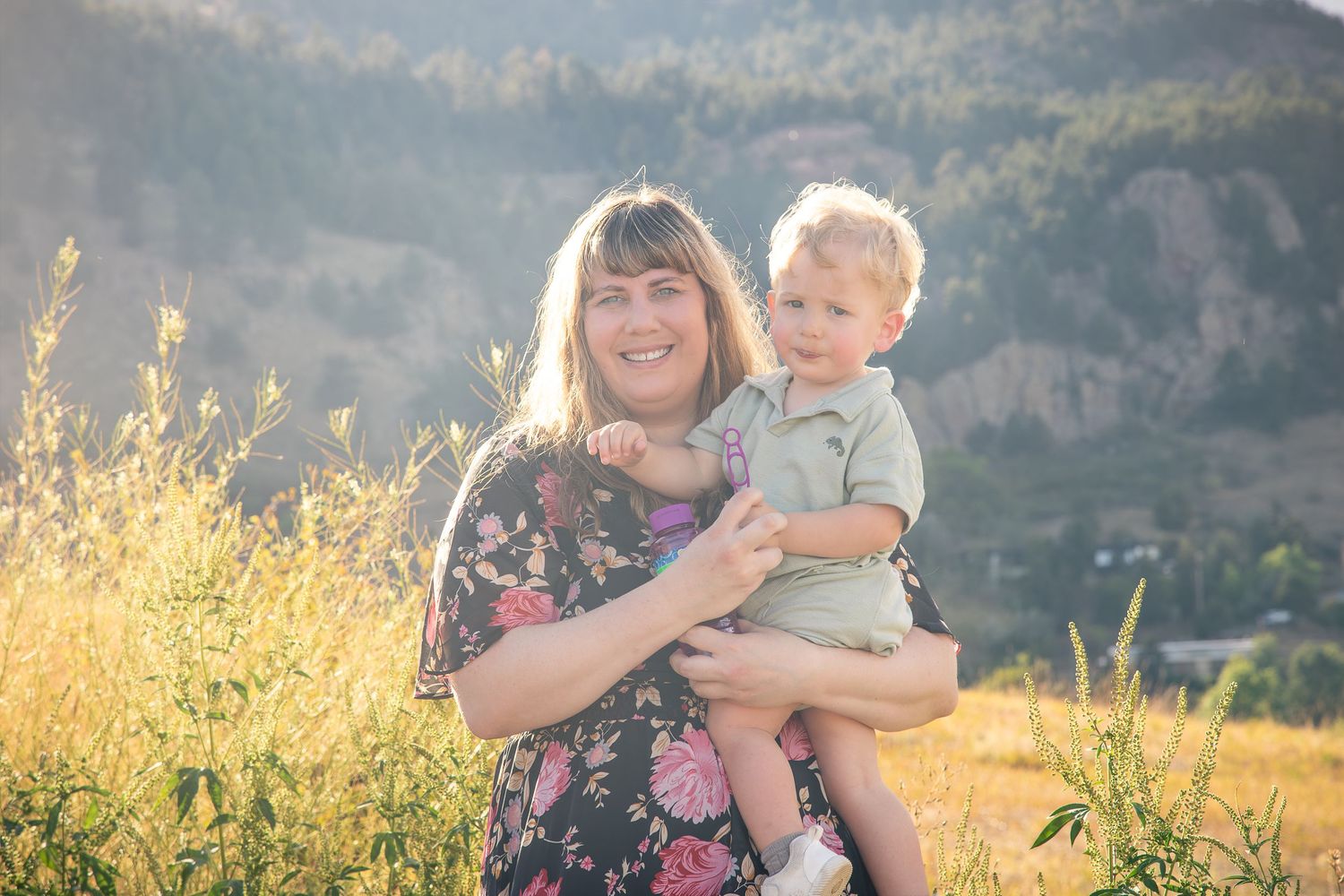 A mother holds her toddler son in a sunny field with mountain backdrop during a golden hour family photo session.