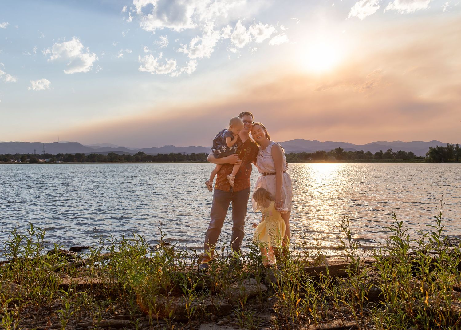 Family posing by a lake at golden sunset with mountains in background, children and parents sharing a warm embrace outdoors.
