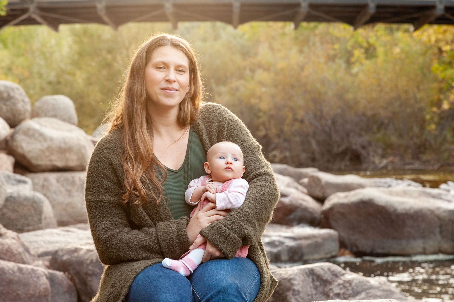 A young mother holds her newborn baby outdoors near a rocky riverbank with autumn foliage in the background.