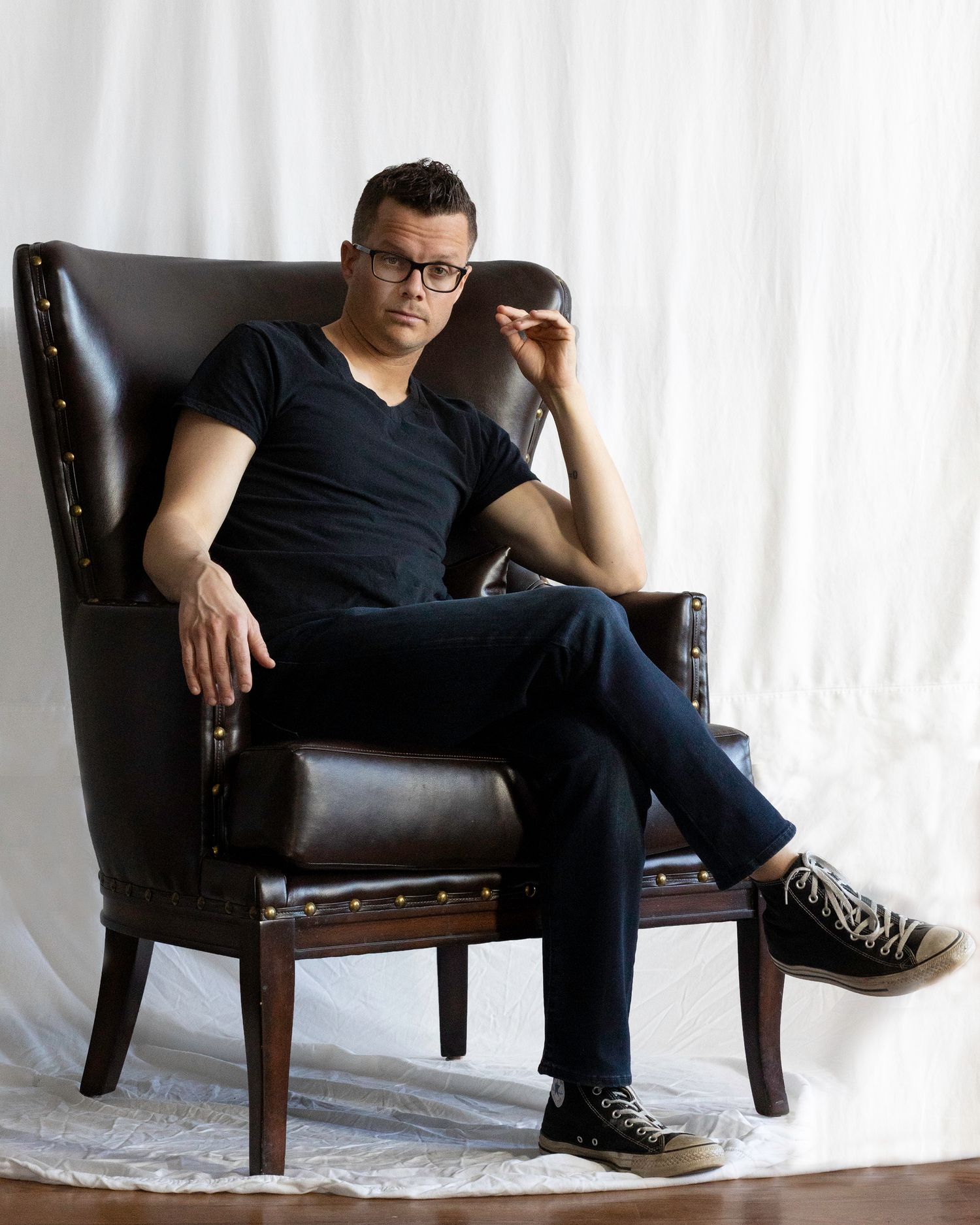Man with glasses sitting in brown leather wingback chair wearing black t-shirt and jeans.