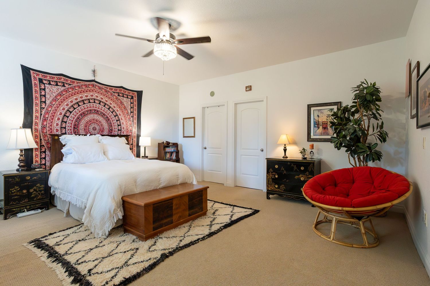 Spacious bedroom with bohemian tapestry, white bedding, red papasan chair, and ceiling fan on beige carpet.
