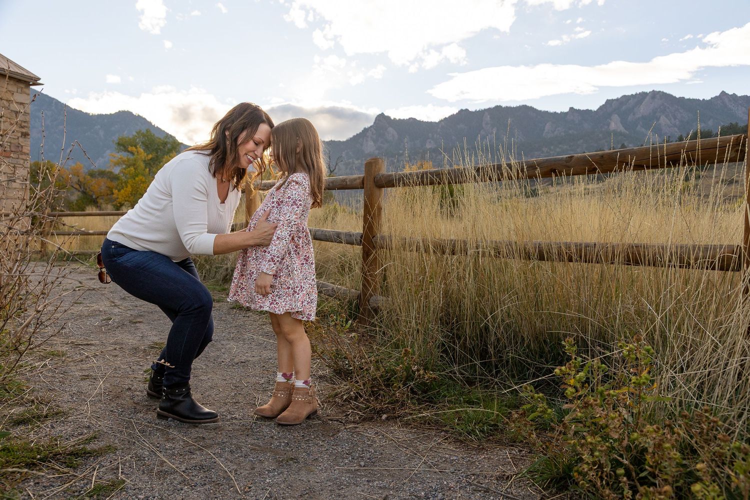 A mother bends down to kiss her young daughter in a floral dress near a wooden fence with mountain views.