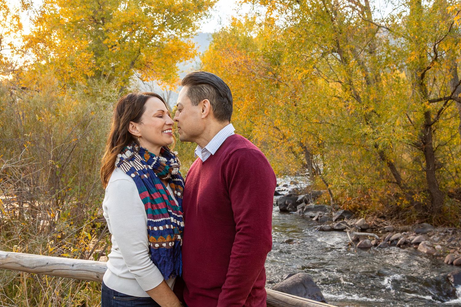 Couple sharing a kiss outdoors in autumn, surrounded by golden foliage and a flowing stream.