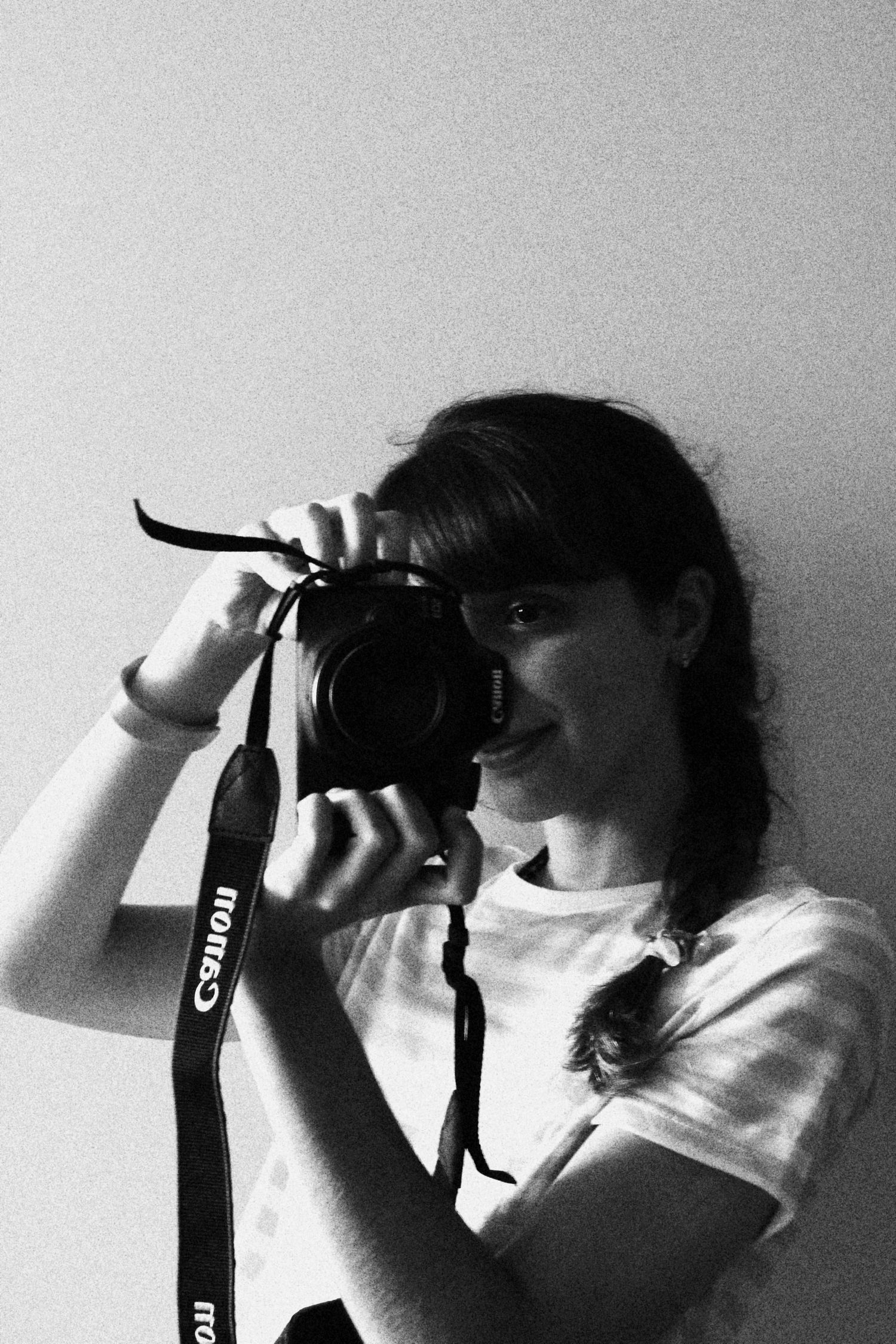 Black and white photo of a young woman taking a self-portrait with a Canon DSLR camera and strap.