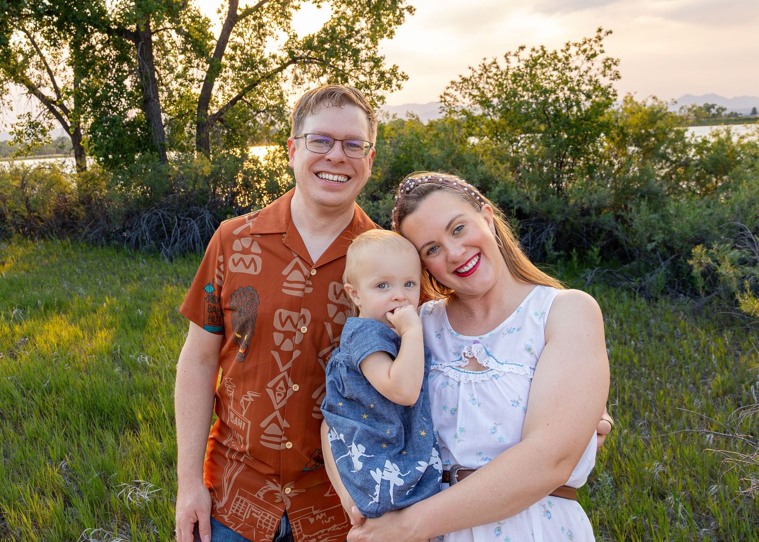 Happy family of three posing outdoors at sunset, parents smiling with baby in a lush green field.
