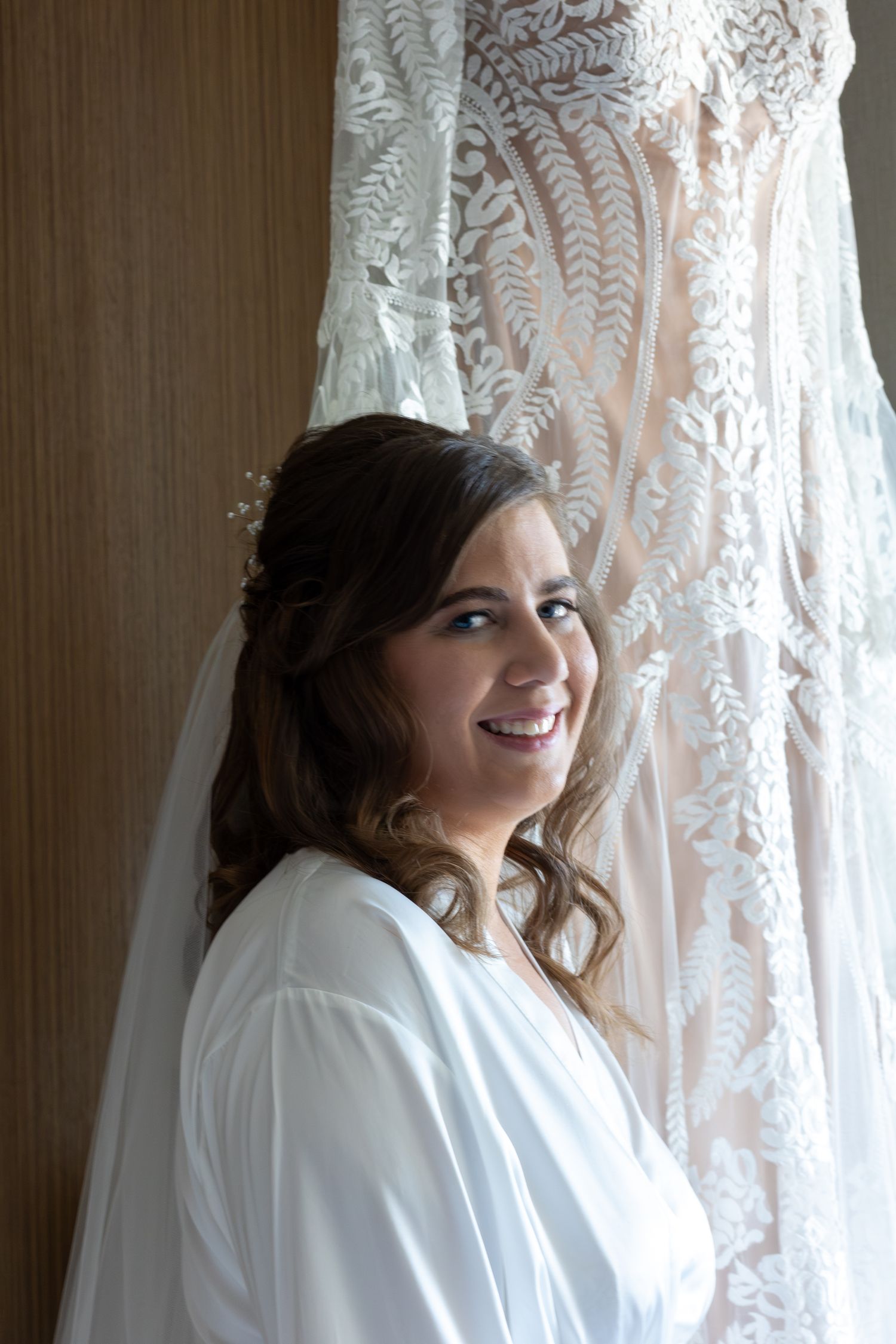 A smiling bride in a white robe posing in front of an elegant lace wedding dress.