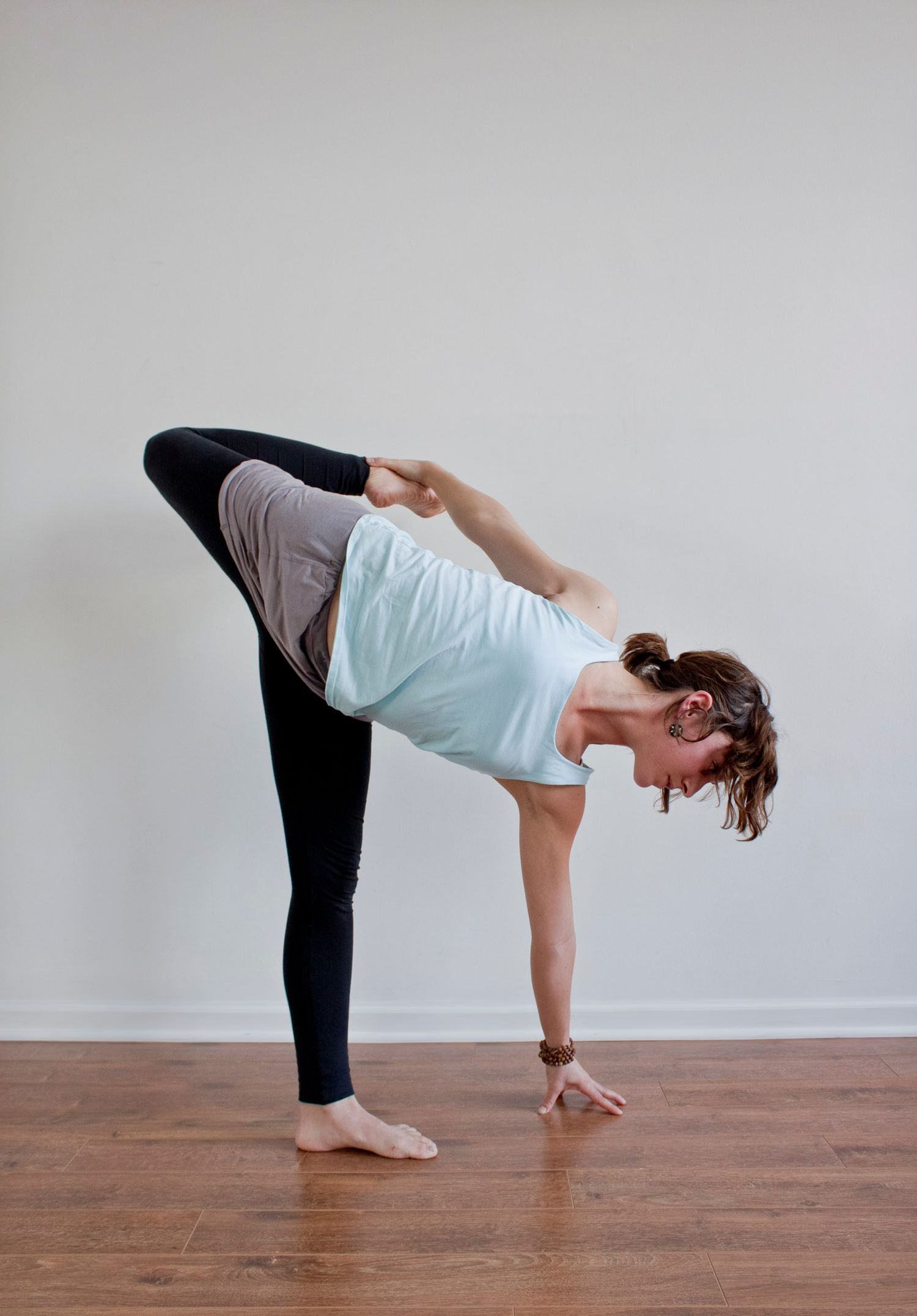 Woman balancing in standing split yoga pose wearing light blue top on hardwood floor.