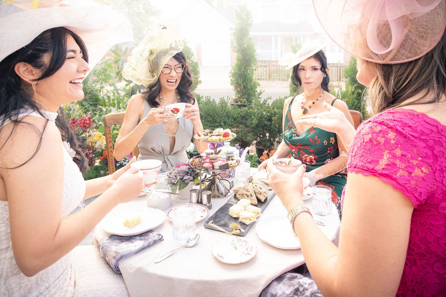 Four women in pastel dresses and fascinator hats laugh together over tea and treats at a garden table during an elegant afternoon tea party.
