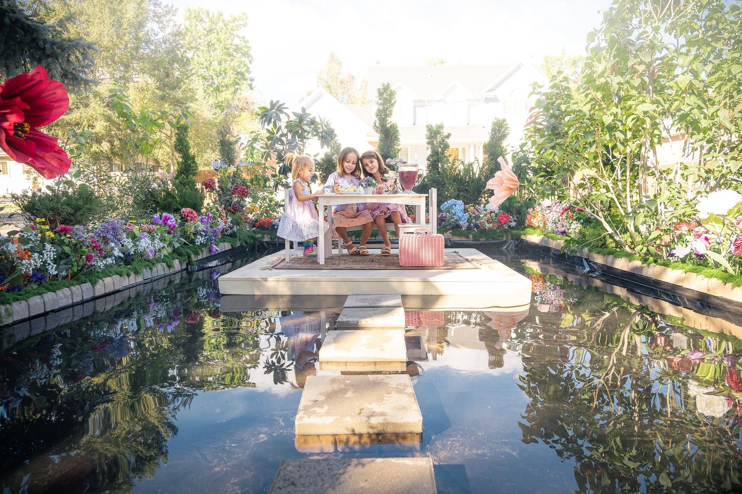 Two children sit at a small pink tea table on a wooden platform surrounded by a reflective pond and colorful flower beds in a garden.