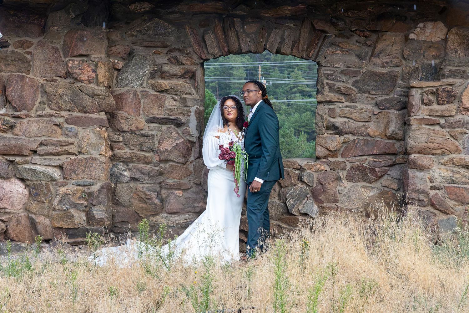 Bride and groom posing in front of a rustic stone wall with an arched window in a field.