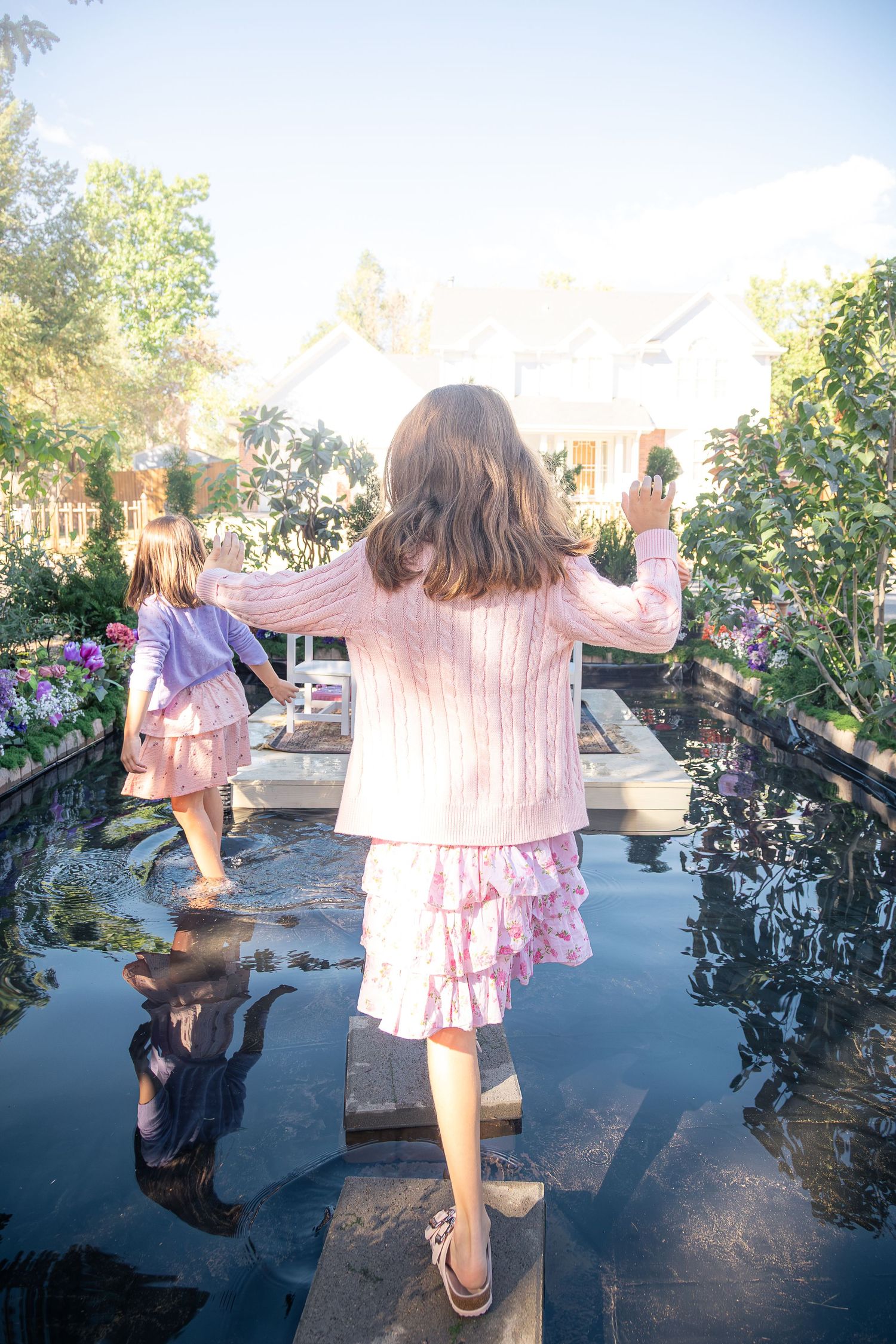 Two young girls in pink outfits walk across stepping stones in a reflective garden pond surrounded by lush flowers and greenery.