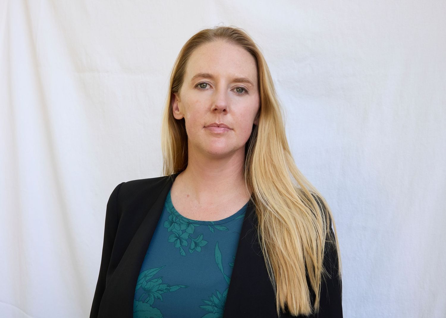 Blonde woman with serious expression in black blazer and teal top against white background.