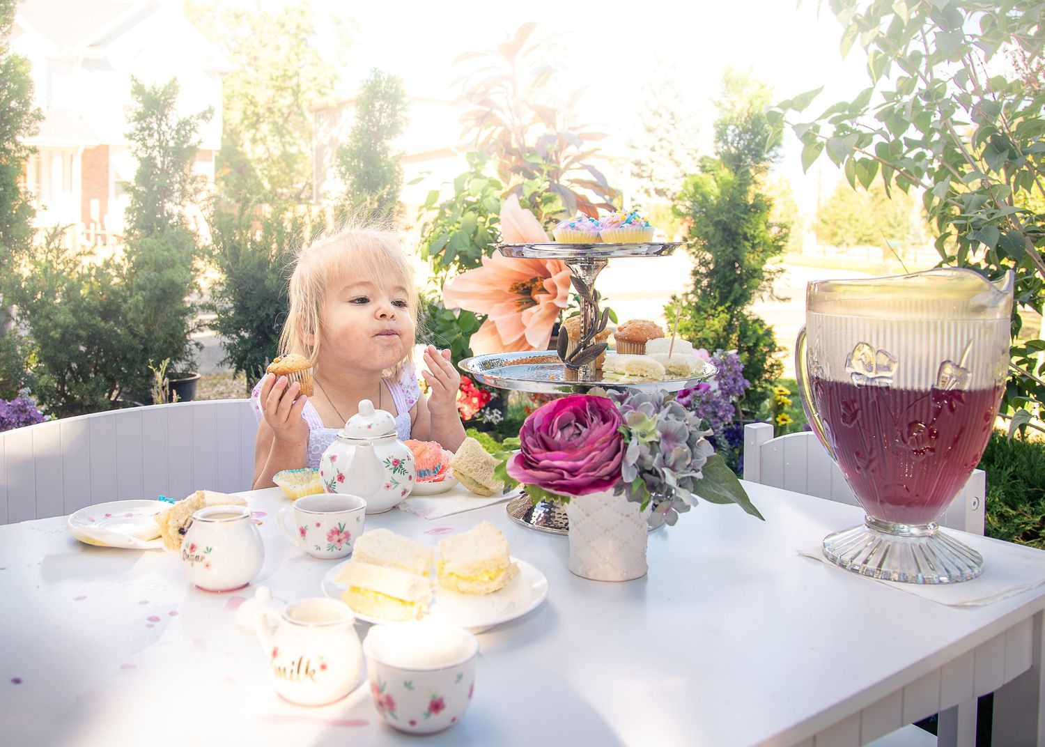 Young blonde girl enjoys a garden tea party at a white table with teapot, cups, sandwiches, pink roses and a tiered stand.
