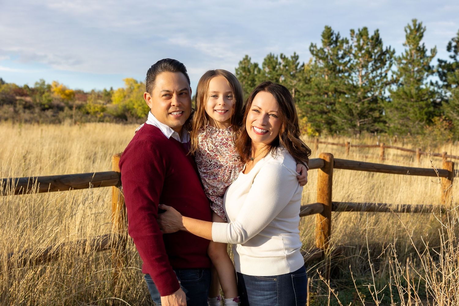 Family of three posing outdoors in a golden field with pine trees and wooden fence in autumn.