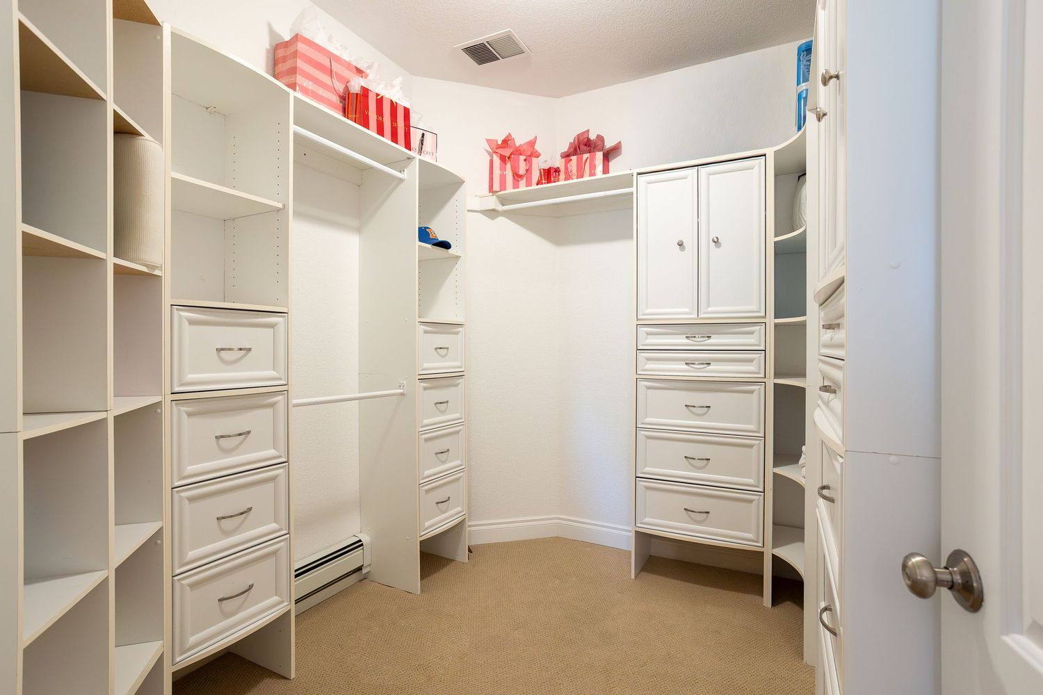 Spacious walk-in closet with white built-in shelving, drawers, and cabinets on beige carpet flooring.