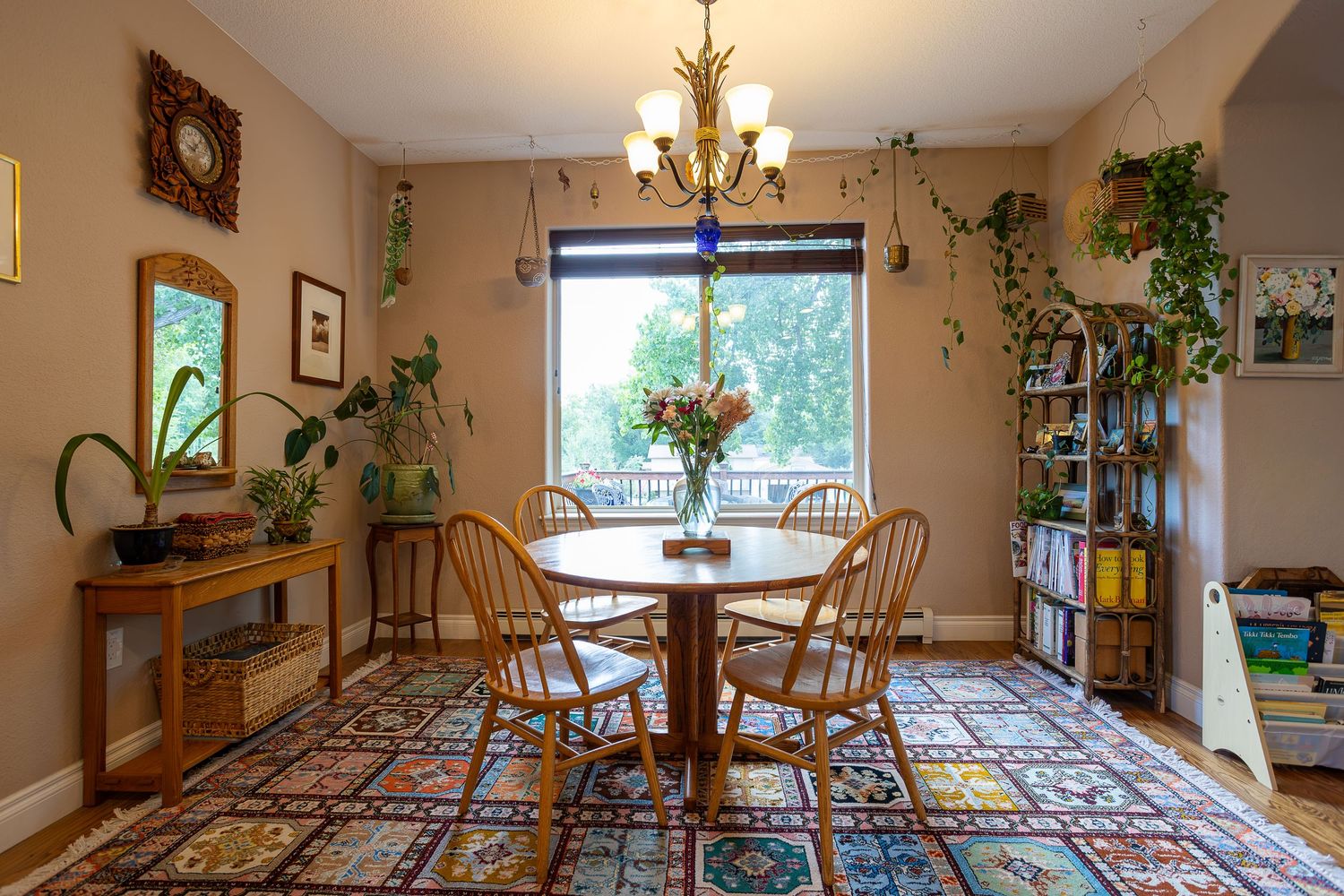 Cozy dining room with round wood table, wicker chairs, colorful rug, chandelier, and lush green plants by a bright window.