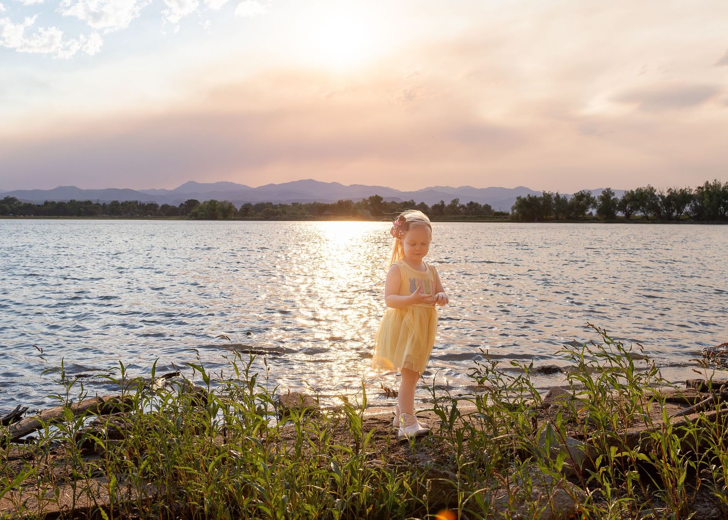 A young girl in a yellow dress stands by a sunlit lake with mountains and trees in the background at sunset.