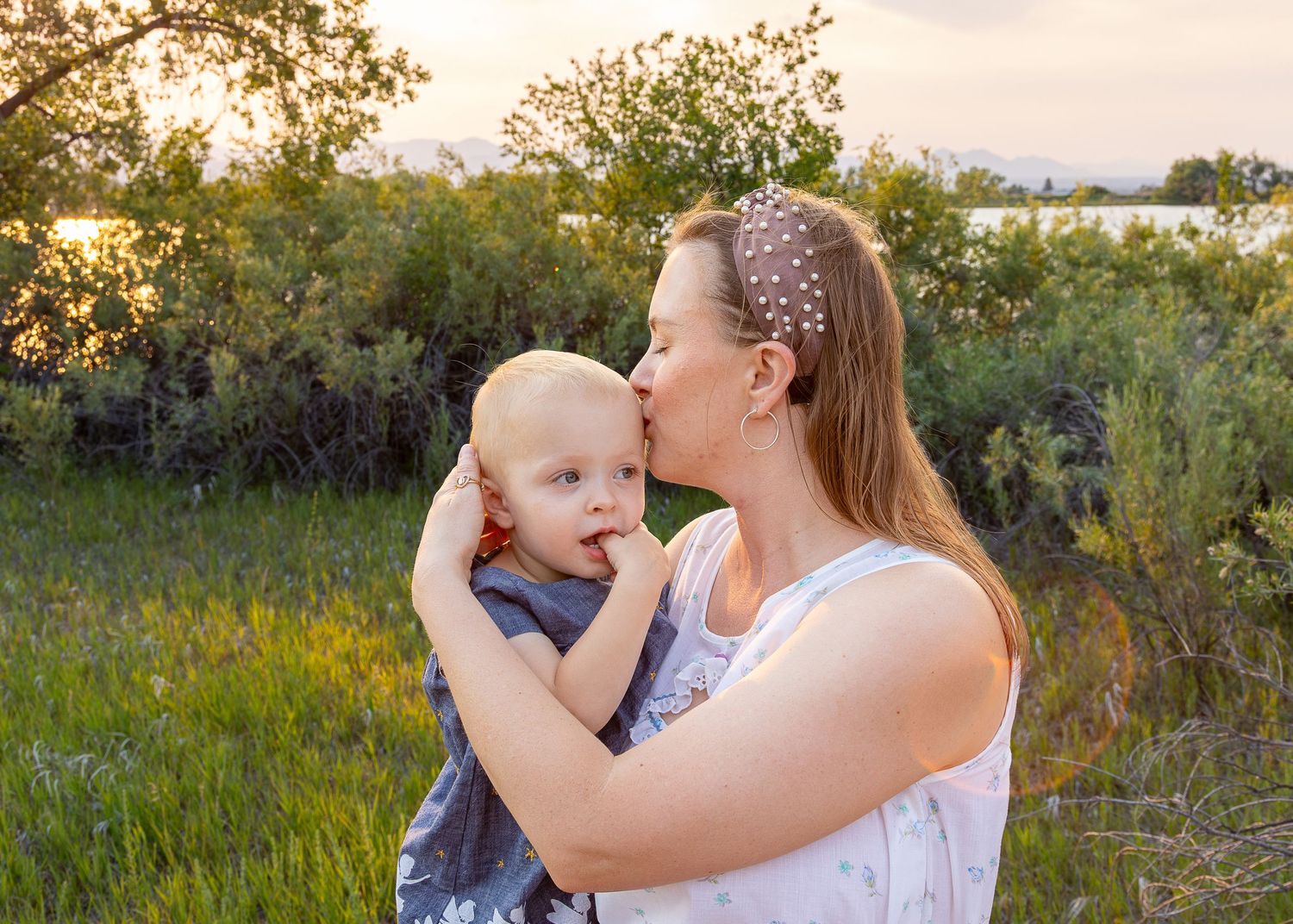 A mother tenderly kisses her young blonde toddler outdoors in a lush green field at golden hour sunset.
